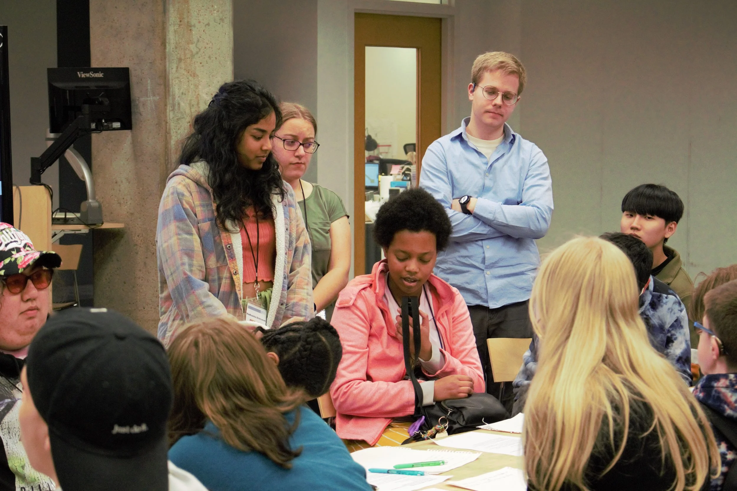 ID: Bella speaks into a small microphone while sitting at a table, surrounded by other participants and mentors both sitting and standing.