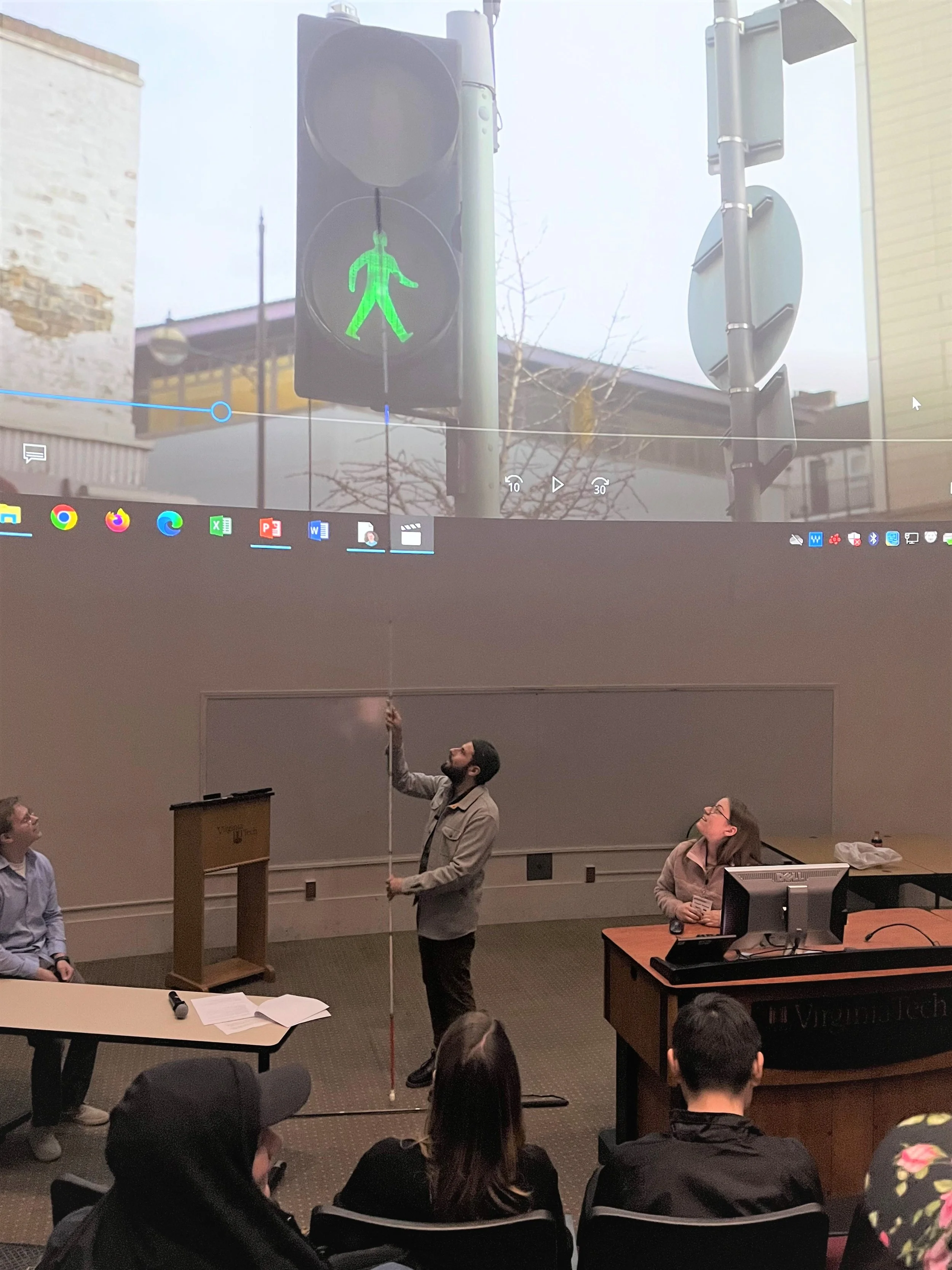 Lecture with artist Carmen Papalia (2023). ID: Carmen stands at the front of a lecture hall with a large projector screen above him. He extends his 50-foot long cane while the audience looks up in awe.