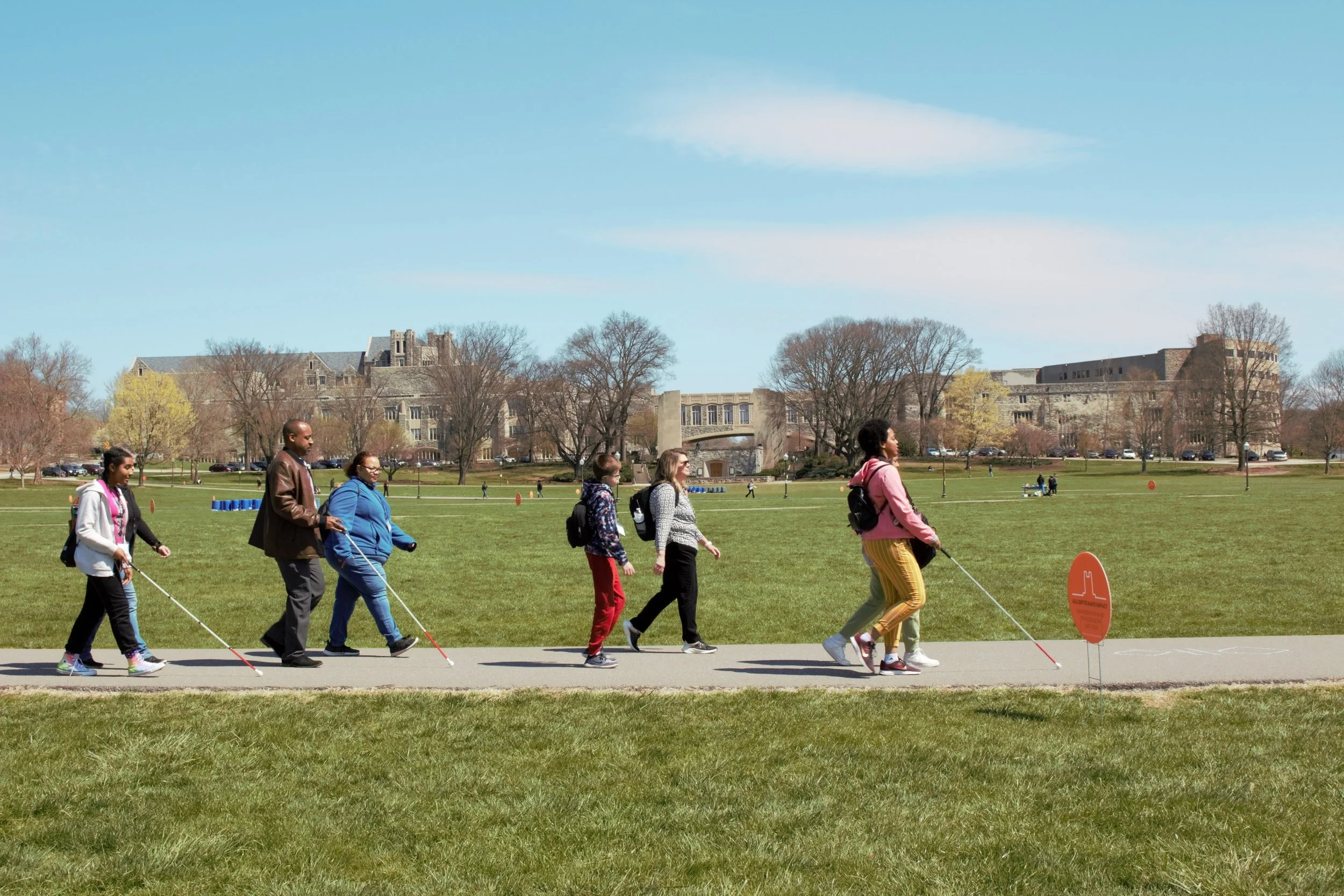 ID: Participants walk with white canes along a paved path on a large grassy field. In the distance is a line of trees and Torgersen Hall.