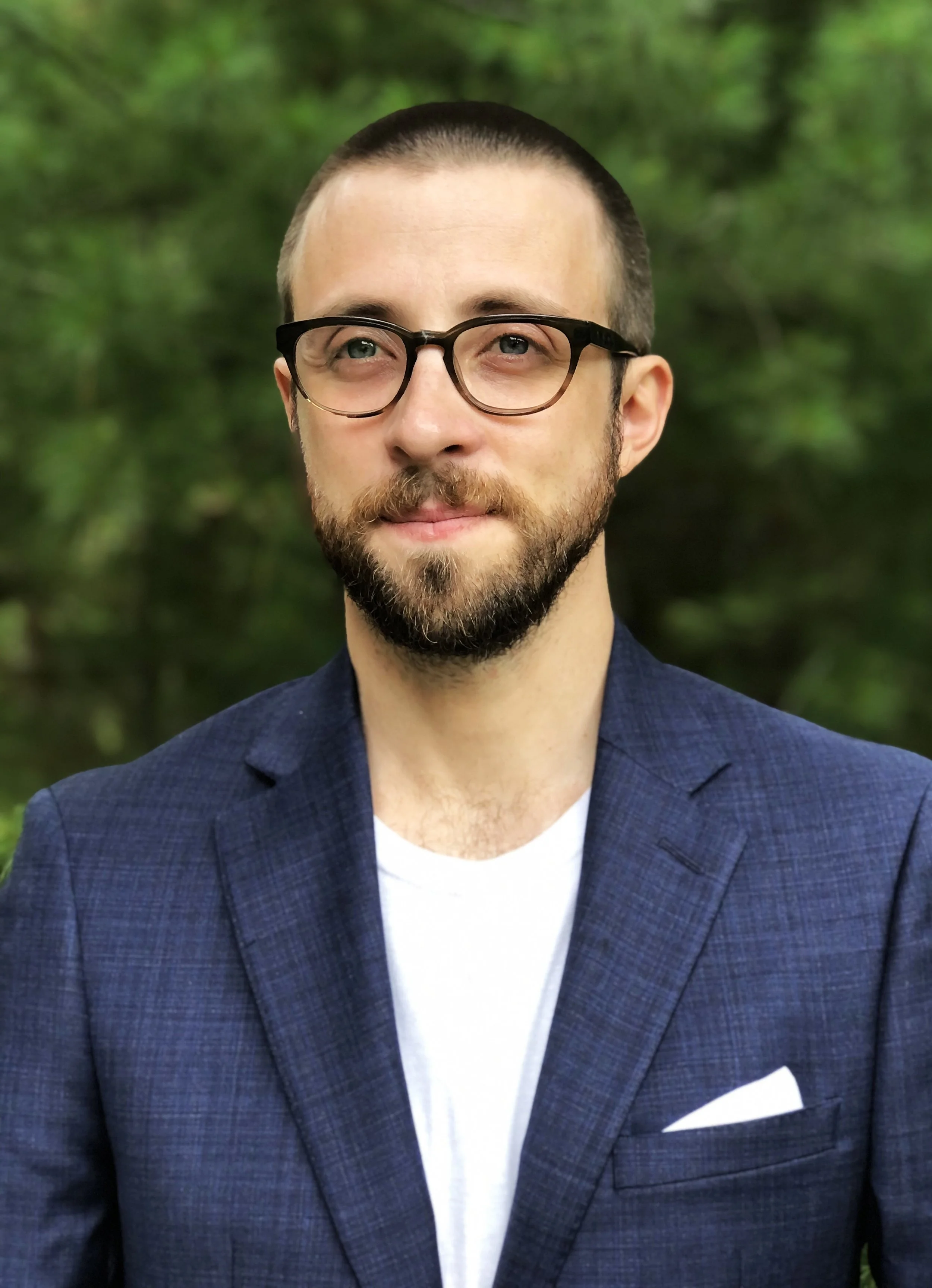 Portrait of a white man with short, dark hair, a beard, black glasses, and wearing a blue blazer over a white shirt.
