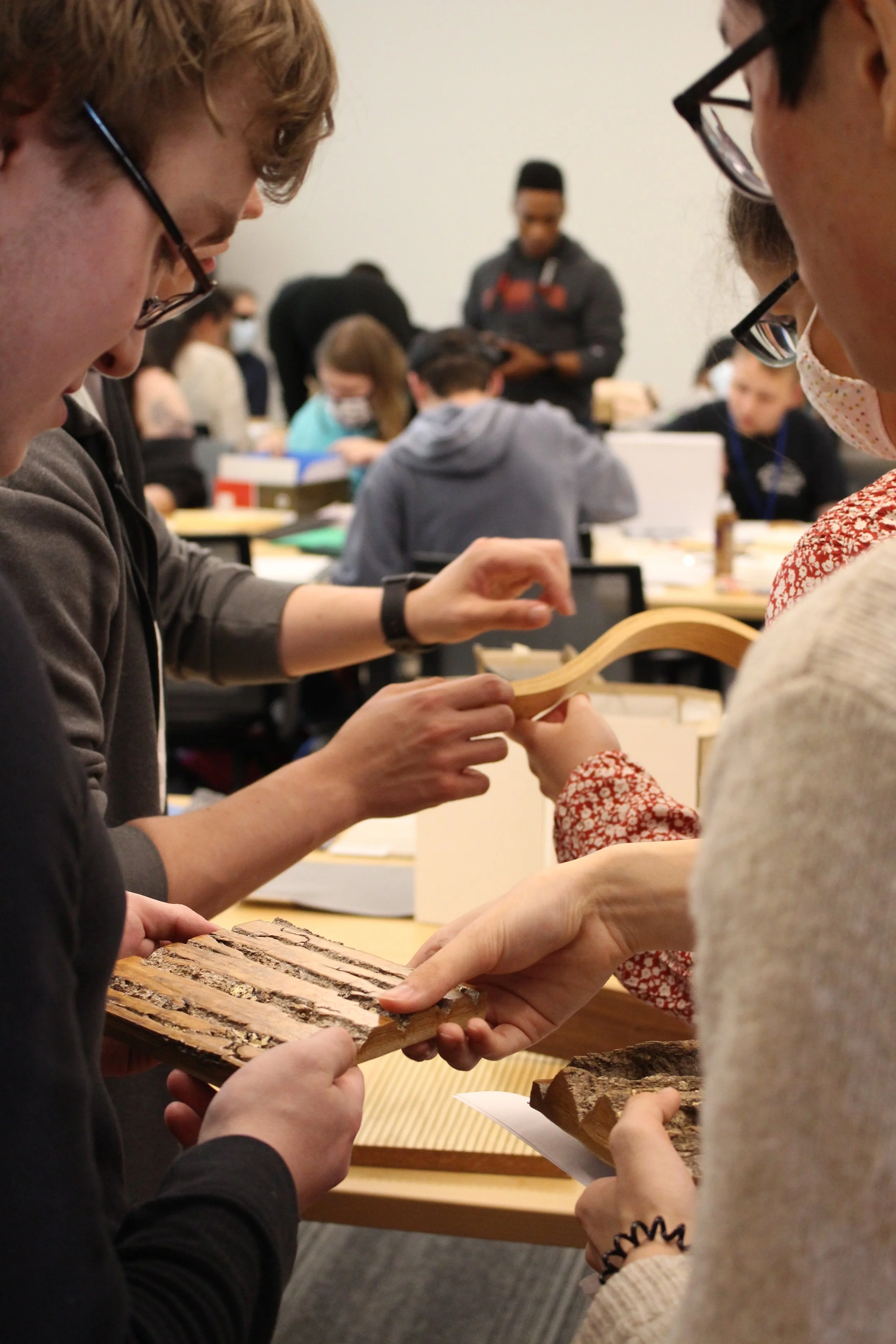 Material explorations with VMDO Architects. ID: Close-up view of hands holding samples of curved wood, with participants sitting at long tables in the background working on models.