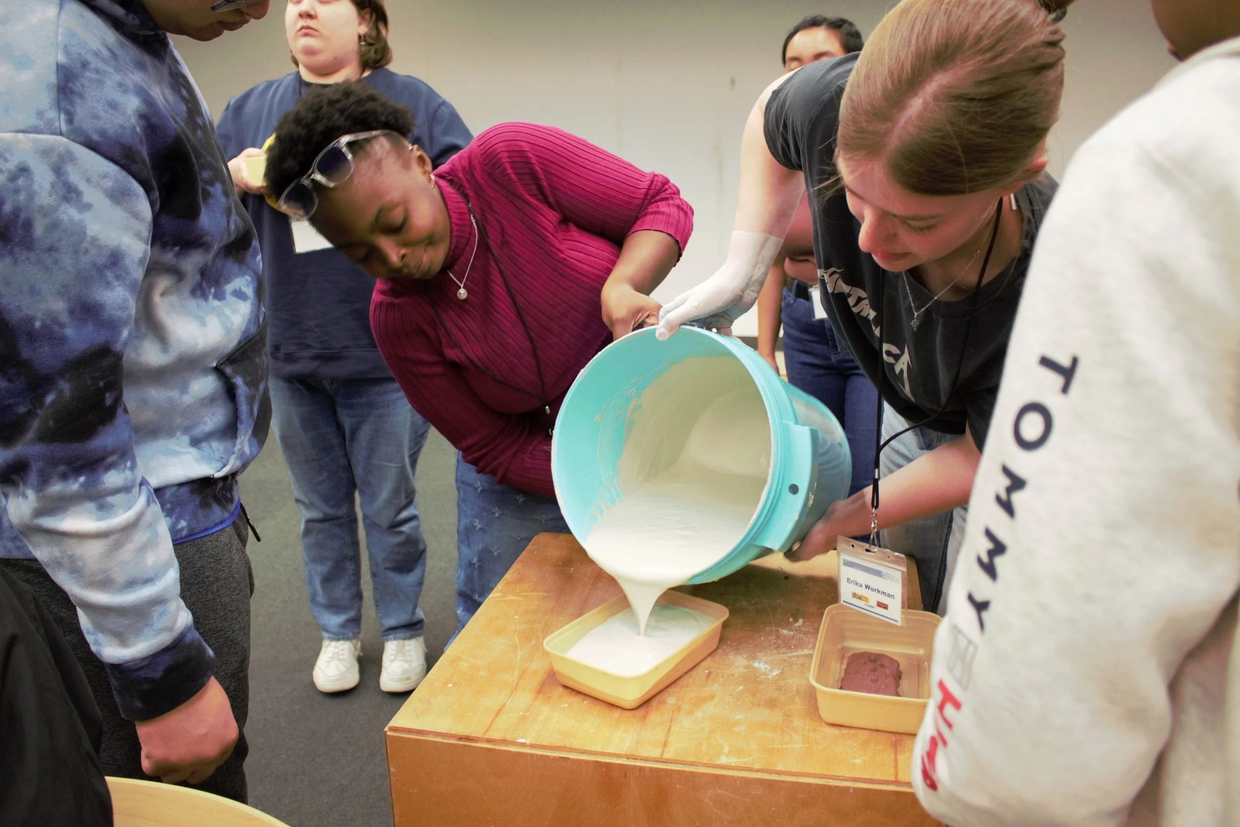Plaster casting activity. ID: Tamia and Erika carefully pour plaster from a large blue bucket into a small Tupperware.