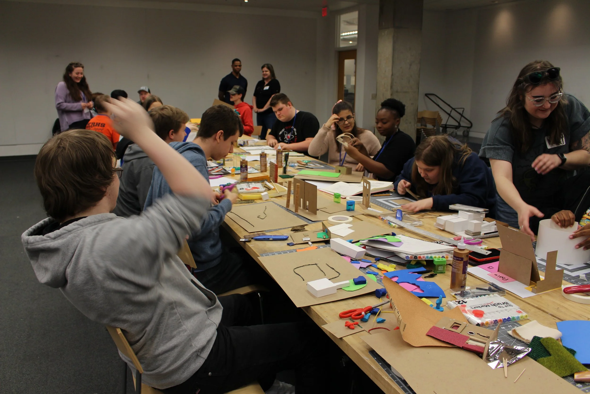 ID: Participants and mentors sit around a long, narrow butcher block table covered in craft supplies and in-progress models.