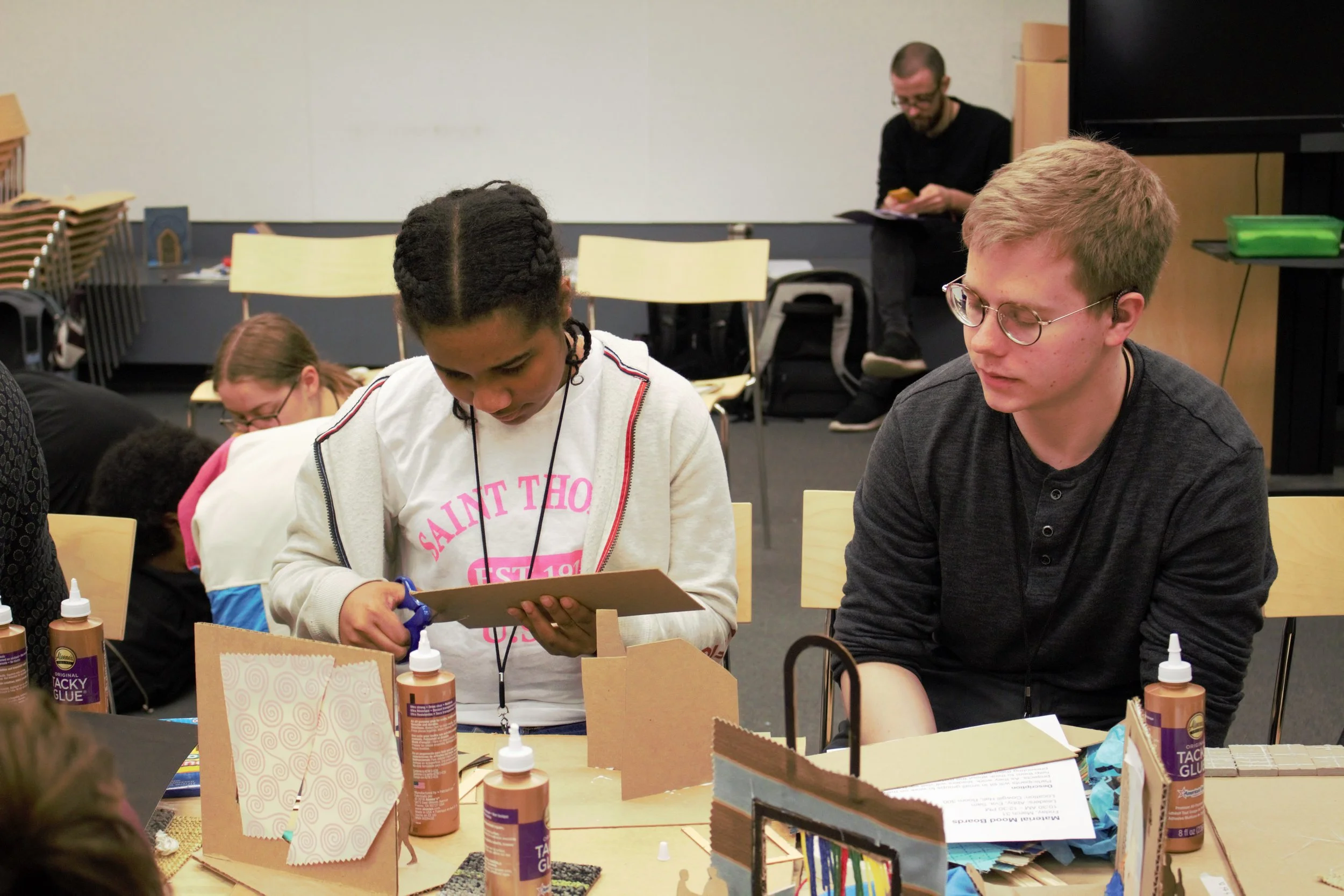 ID: Front-facing view of Jordyn and Matthew sitting at a table while Jordyn cuts a piece of cardboard with scissors.
