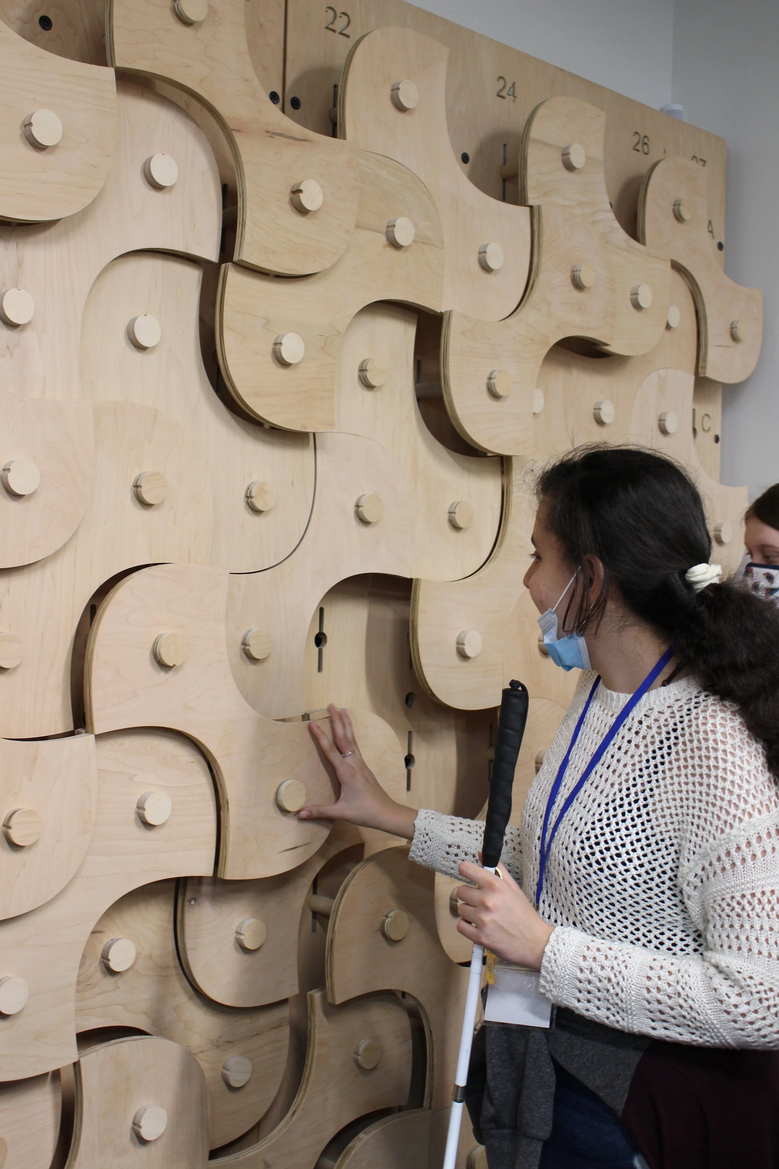 Tour of student design-build projects in Virginia Tech's new Creativity & Innovation District building. ID: Mariam touches a decorative wall composed of many curving wood panels.