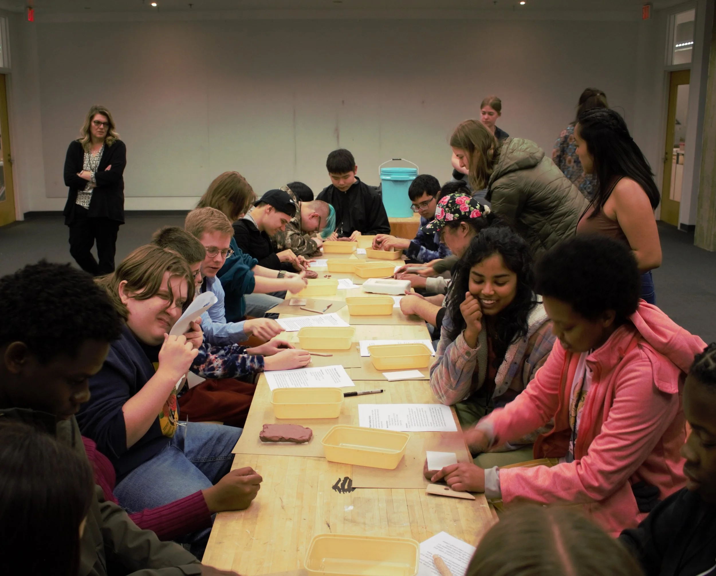 ID: Participants and mentors sit at a long butcher block table with tan Tupperwares and white instruction packets in front of them.