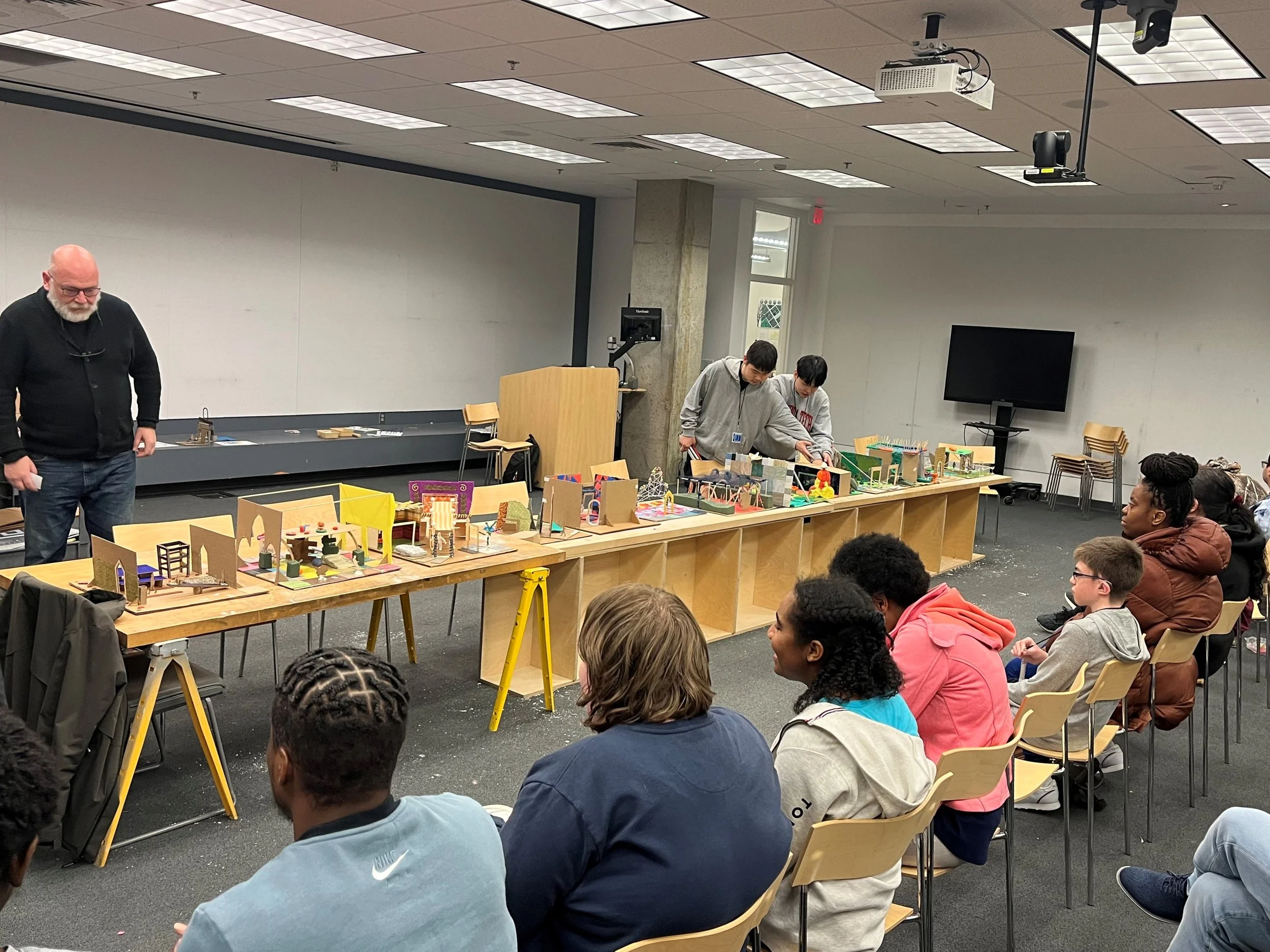 Participants present their final models for a group discussion. ID: An audience faces a long butcher block table covered in the participants' models.