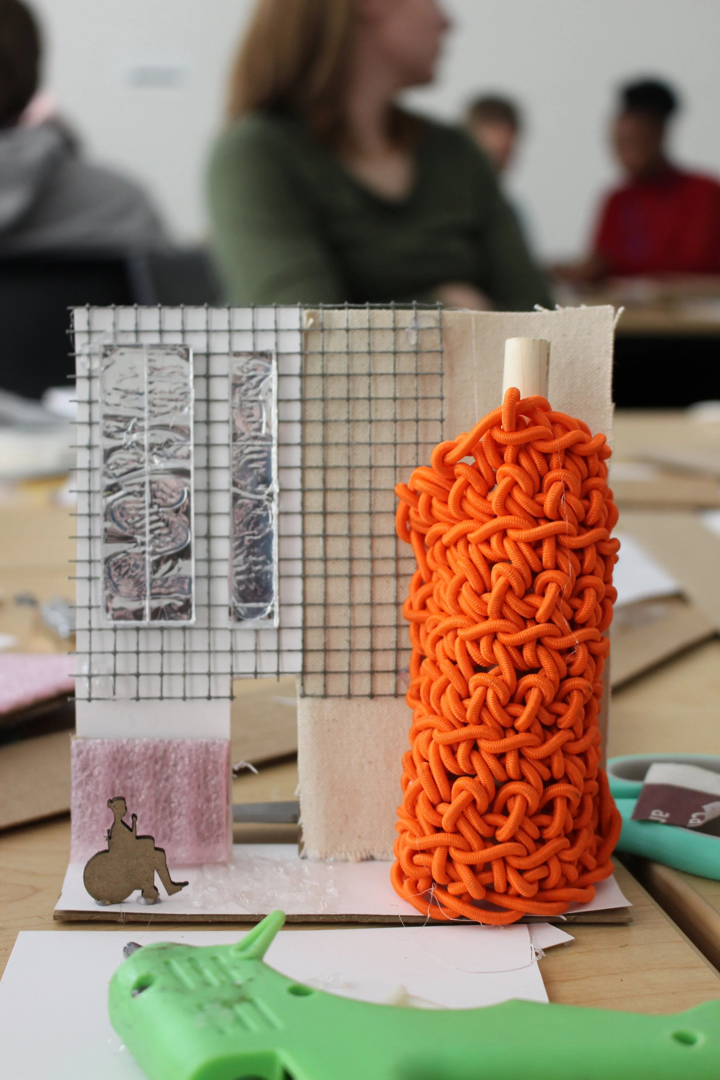 Facade materiality activity. ID: Close-up photo of a participants' facade model, a flat piece of cardboard decorated with metal lath and fabric. A large, bright orange jumble of lace supposedly represents a tree. A wheelchair user cut from cardboard 