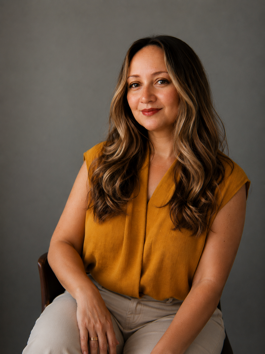 A woman with shoulder-length wavy brown hair, wearing a rust-colored turtleneck, resting her head on her hand, smiling gently at the camera.