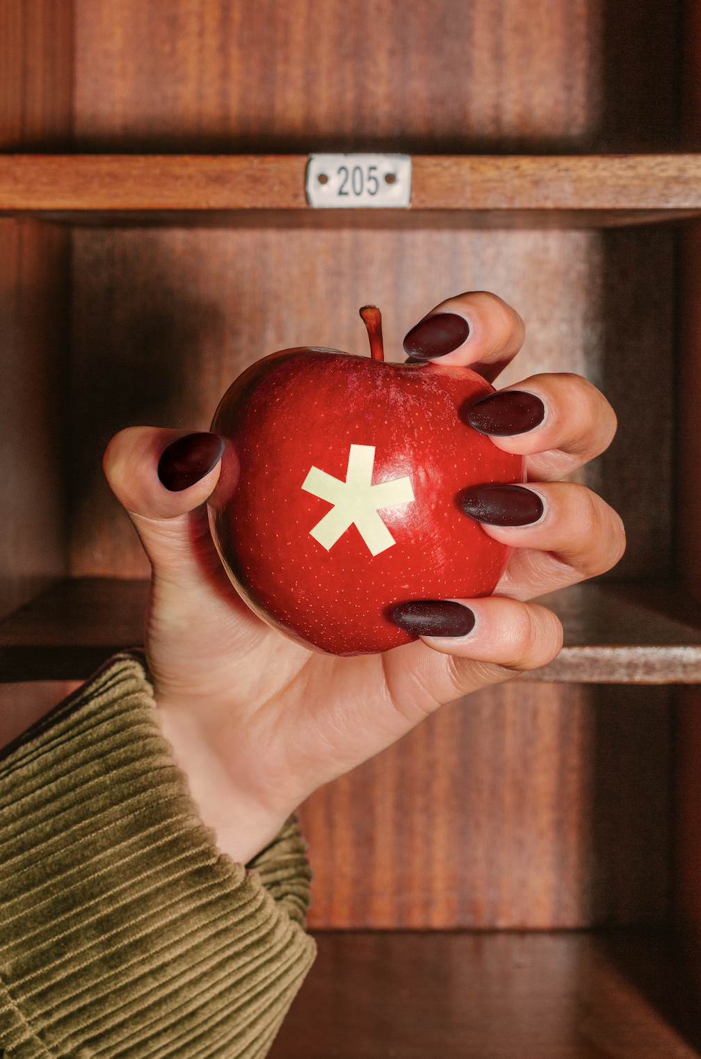 A hand with dark red painted nails holding a red apple with a white asterisk sticker on it in front of a wooden cubby labeled '205'.