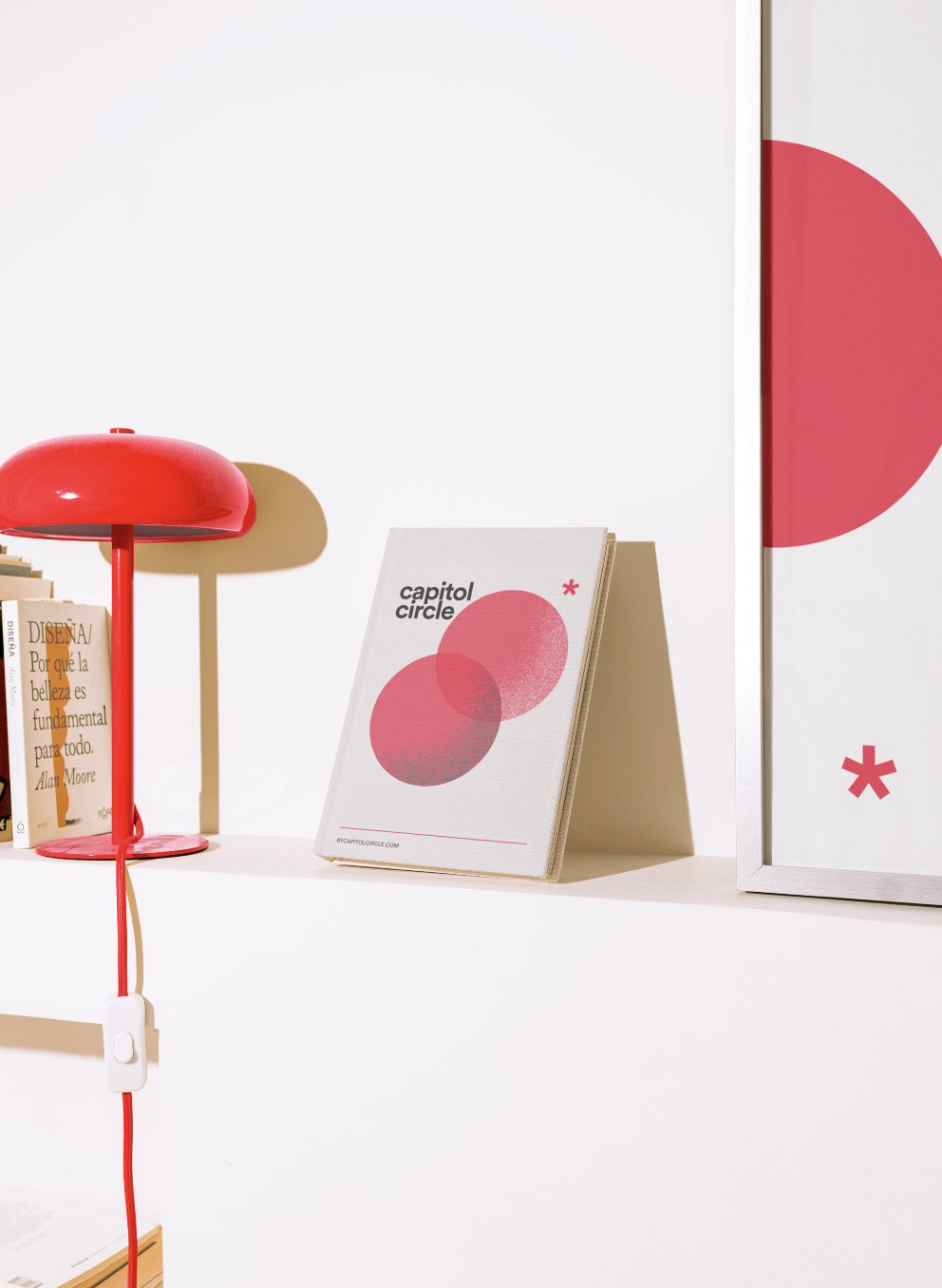 White desk with a red lamp, a row of books, and a triangular display stand with a booklet titled 'Capitol Circle' featuring pink circles on the cover. There is a framed artwork with pink circles and a small pink asterisk.