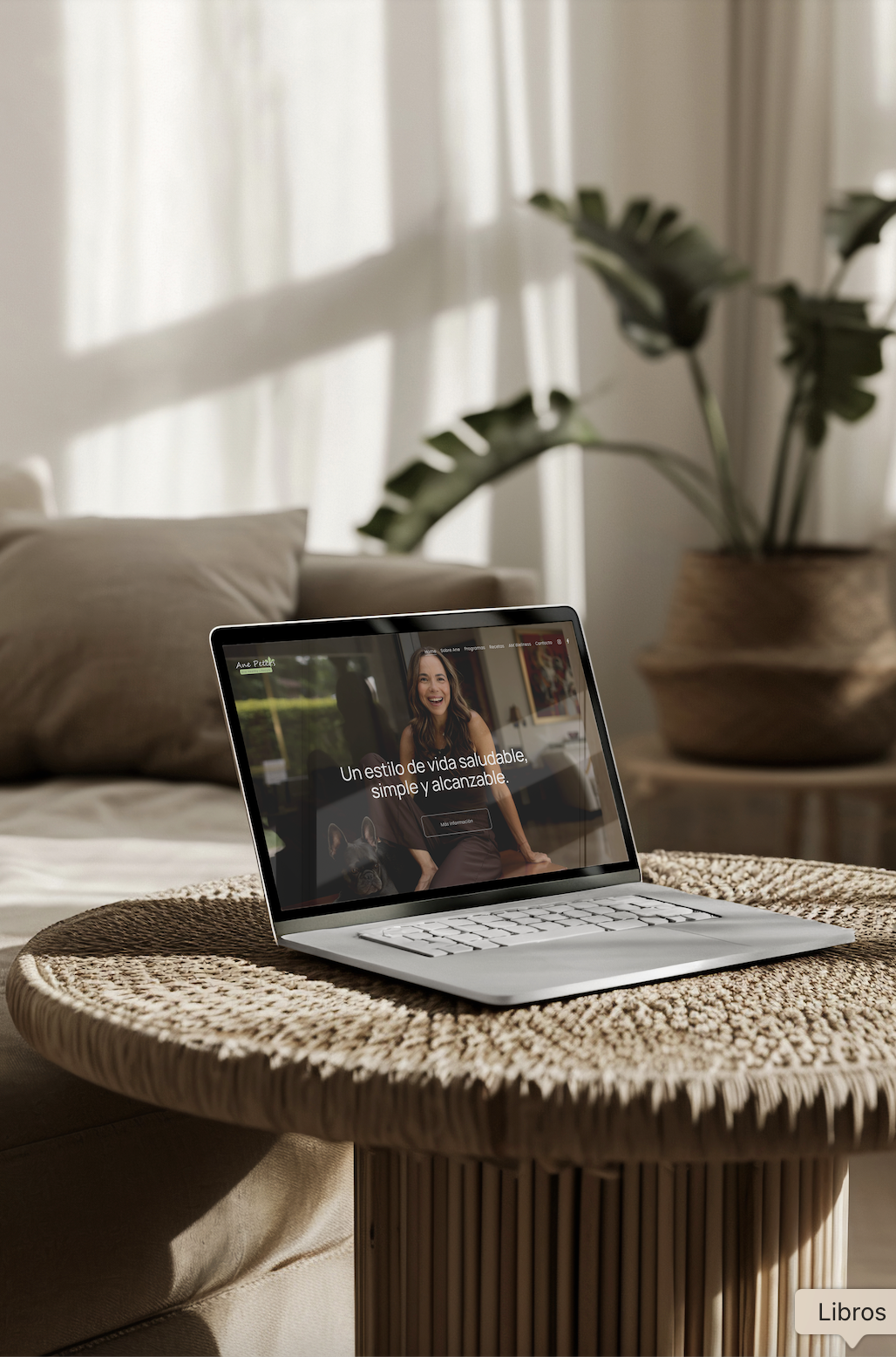 A laptop on a wicker table displaying a woman smiling with a dog on a website, in a cozy room with beige pillows and a large potted plant in the background.