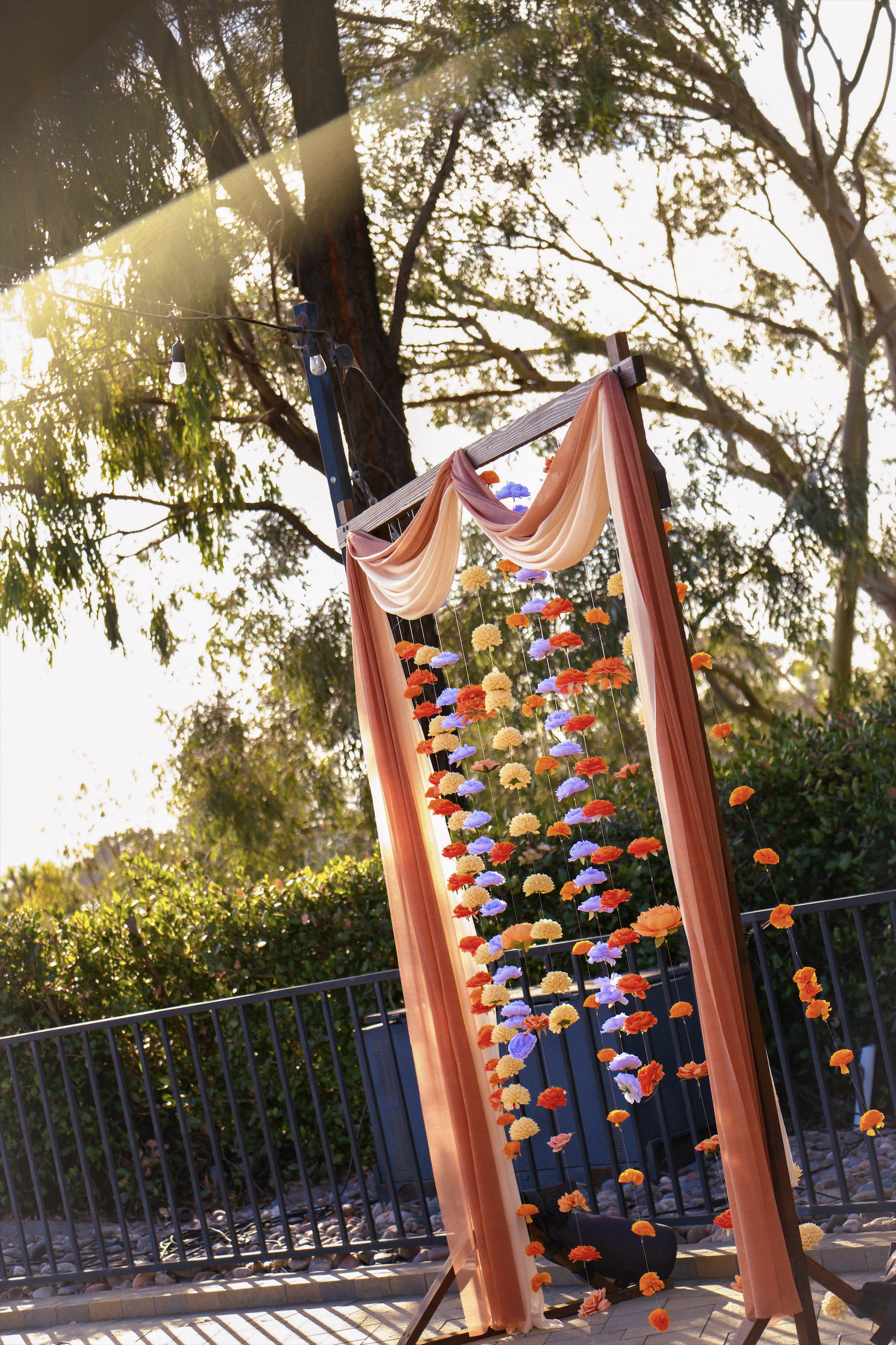 Outdoor floral display with multicolored flowers hanging from a wooden frame, surrounded by trees and sunlight.