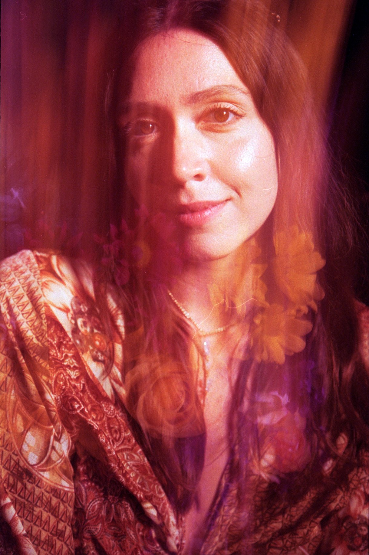 A woman with long brown hair and a patterned blouse, smiling softly, with pink and orange light reflections on her face and clothing.