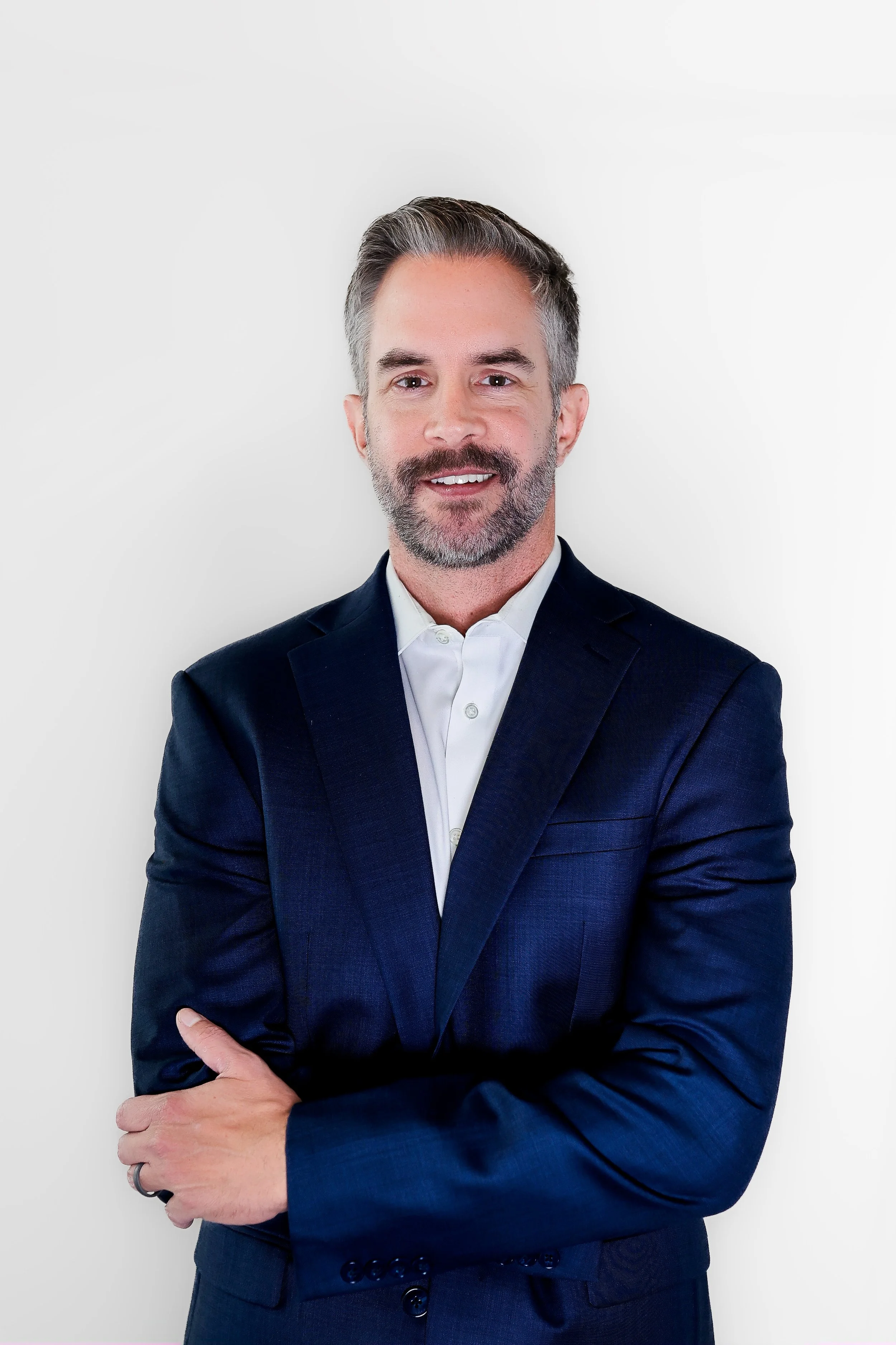 A middle-aged man with short gray hair and a beard, wearing a navy blue suit and white dress shirt, standing against a plain white background.