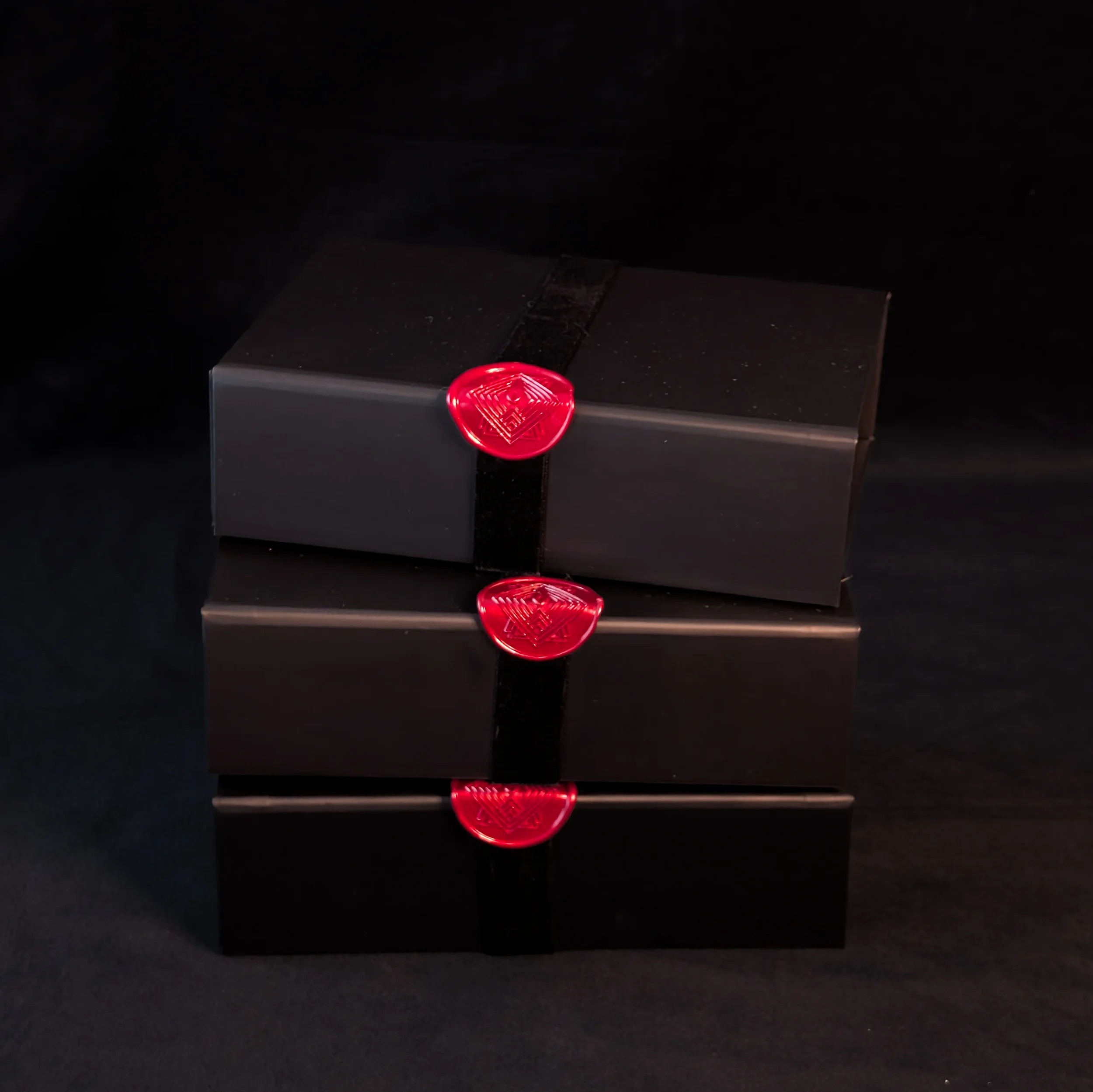 A stack of three black gift set boxes, each closed with a black velvet ribbon and a red wax seal. The image is dark and moody.