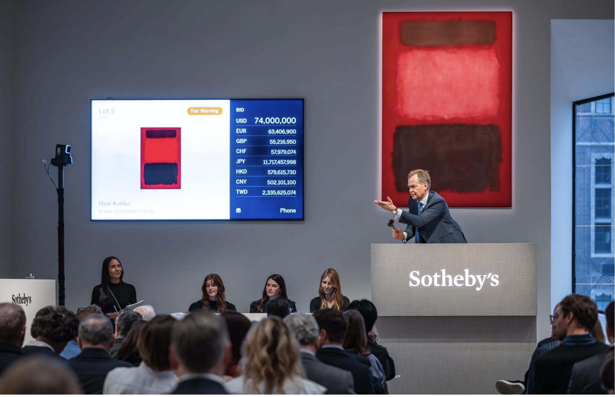 Auction scene at Sotheby's with a man in a suit presenting a piece of art titled 'Brown and Blacks on Reds' by Mark Rothko, while a digital screen displays bidding information including a bid of $74,000,000 in USD and various other currency amounts. Several people are seated, watching the auction.