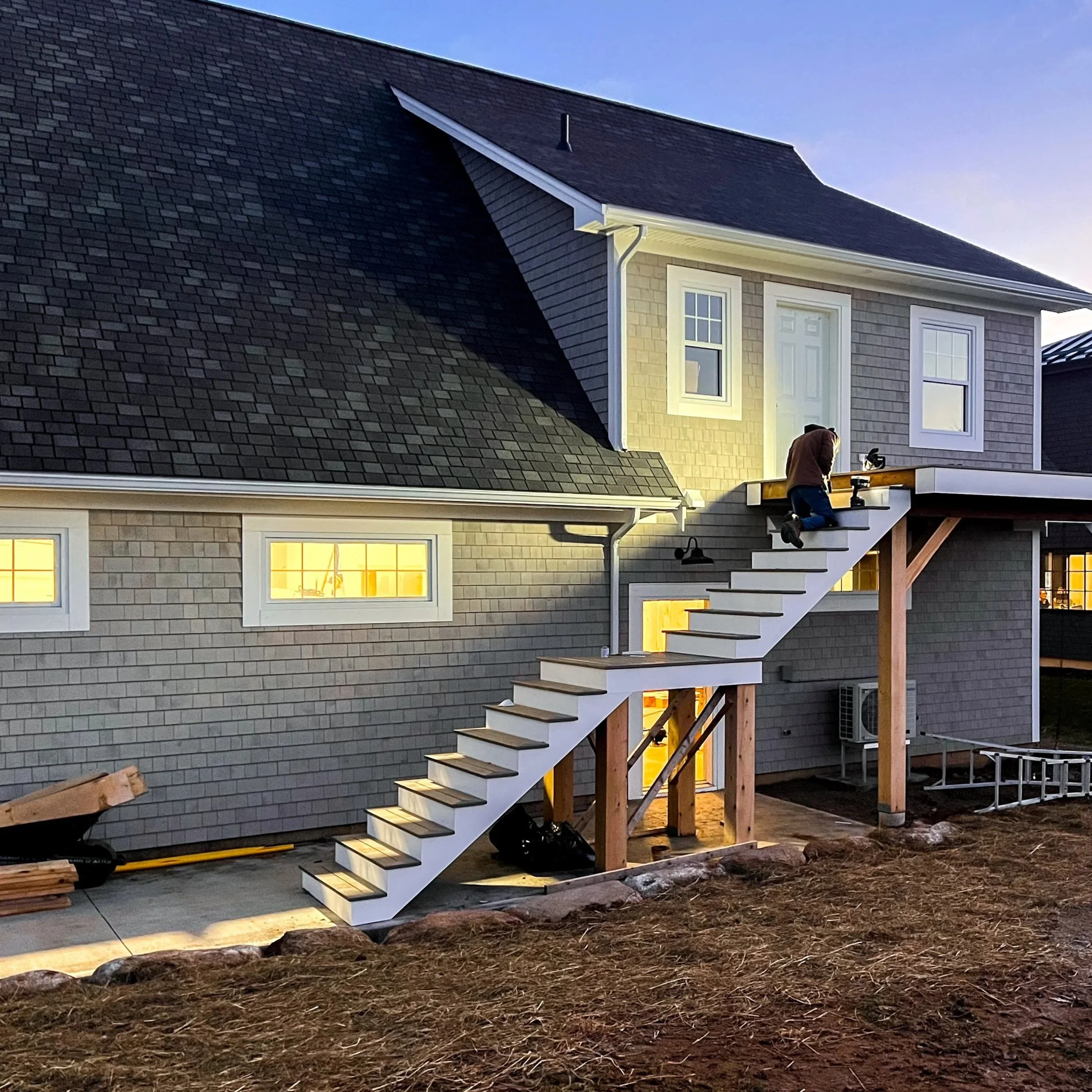A person working on installing a staircase outside a house with gray siding and black shingles, illuminated by exterior lighting at dusk.