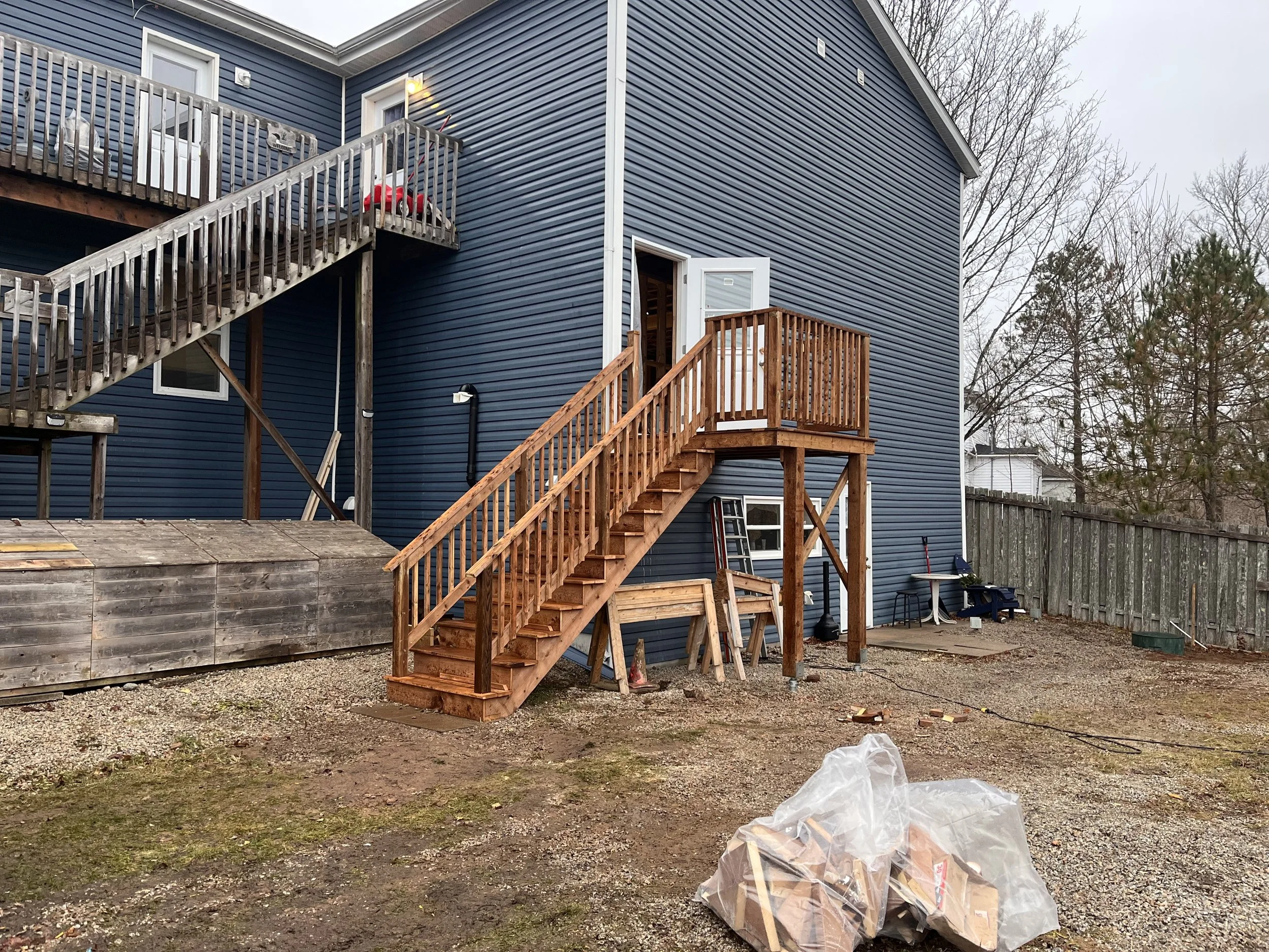 Backyard construction scene with a newly built wooden staircase leading to an open door on a blue house. The backyard is unfinished with dirt, gravel, and construction materials, including a plastic bag of wood scraps and a wooden deck.