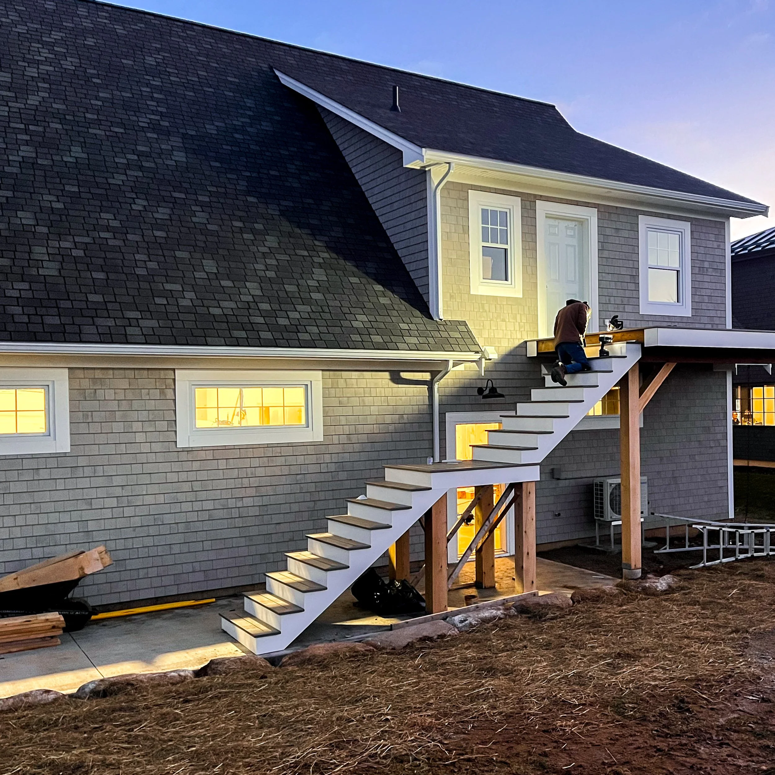 New construction of house in East River Nova Scotia with carpenter finishing a staircase.  The house has grey siding, large windows with warm indoor lighting, and a sloped roof with dark shingles.