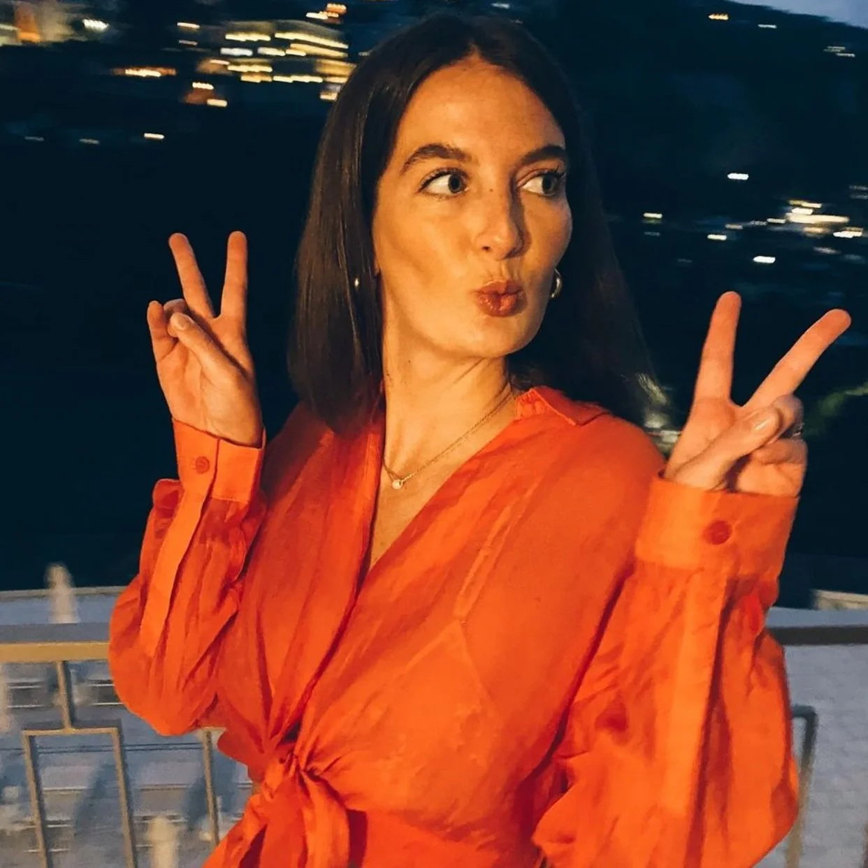 Woman in red blouse making peace signs with both hands, blurred night cityscape background.
