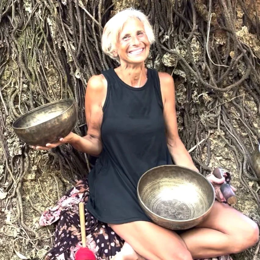 Woman smiling while sitting in nature, holding two singing bowls with roots in the background.