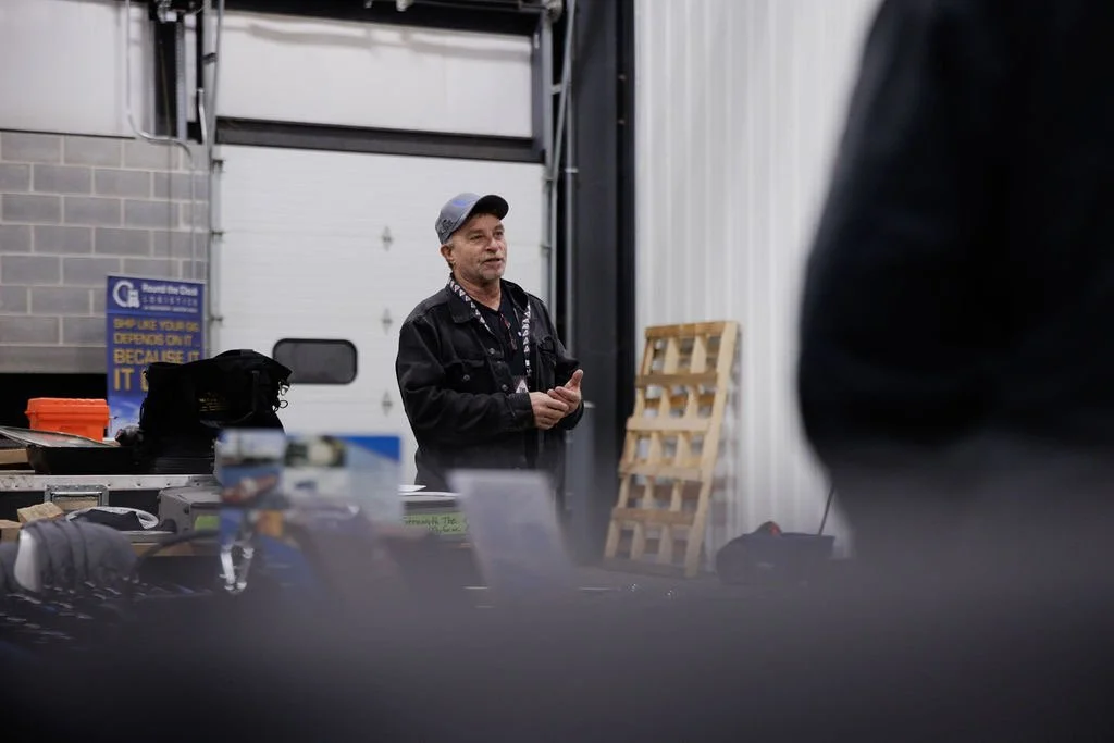 A man with a gray baseball cap and black jacket speaking indoors, with a white garage door in the background and industrial equipment around him.