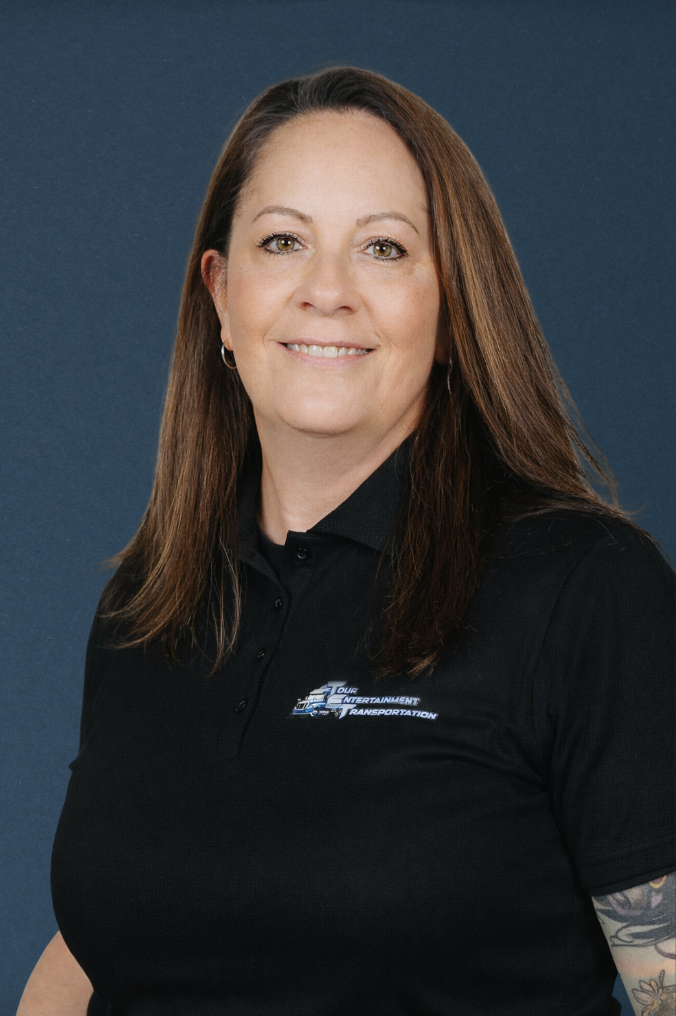 A woman with shoulder-length brown hair, wearing a black polo shirt with a logo on it, smiling against a blue background.
