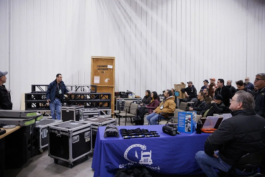 A group of people attending a presentation or training session in a large indoor space, with equipment and a blue tablecloth with a forklift logo on a table in the foreground.