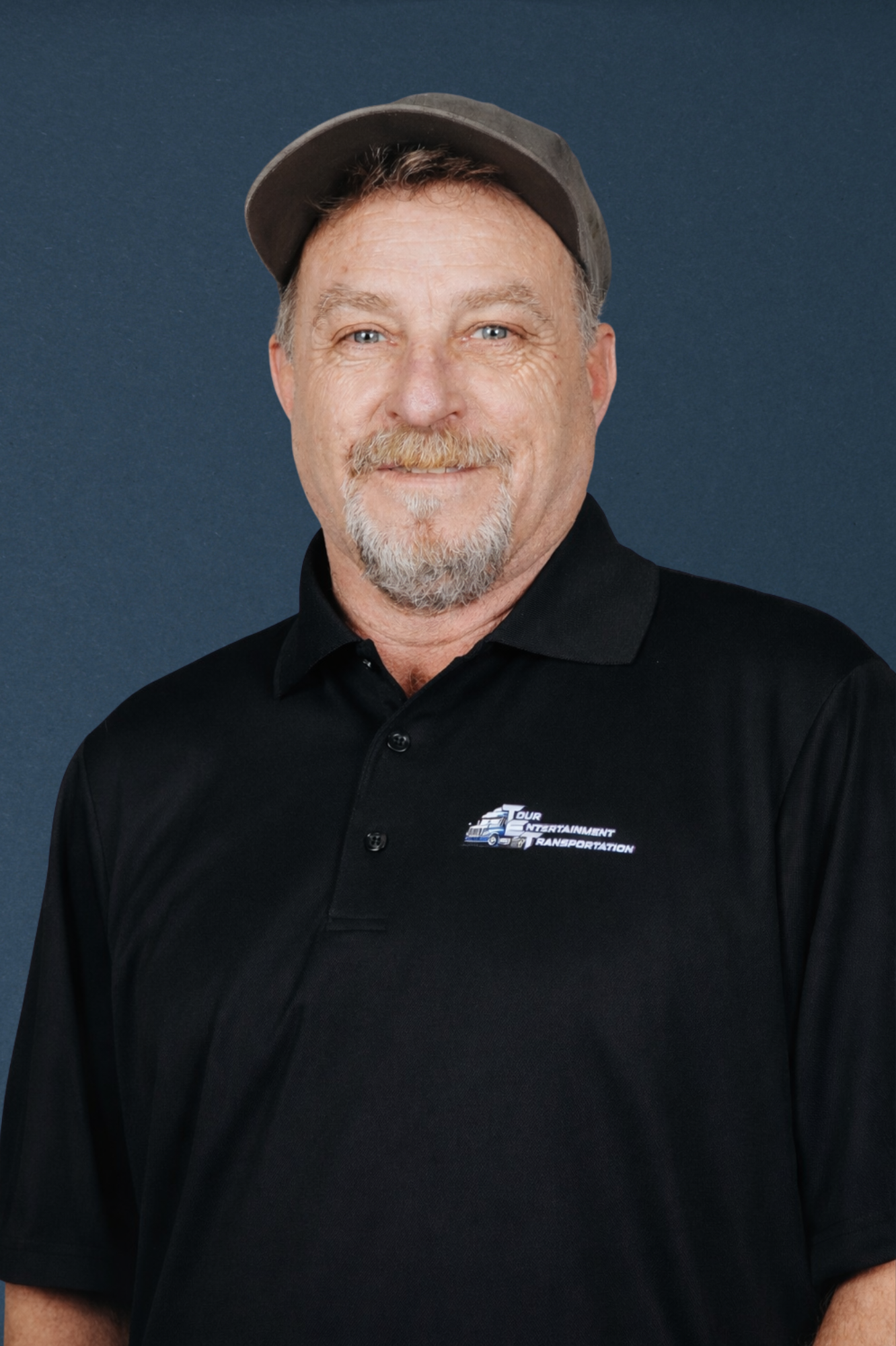 Portrait of a middle-aged man with a goatee, wearing a black polo shirt with a logo and a gray cap, standing against a dark blue background.