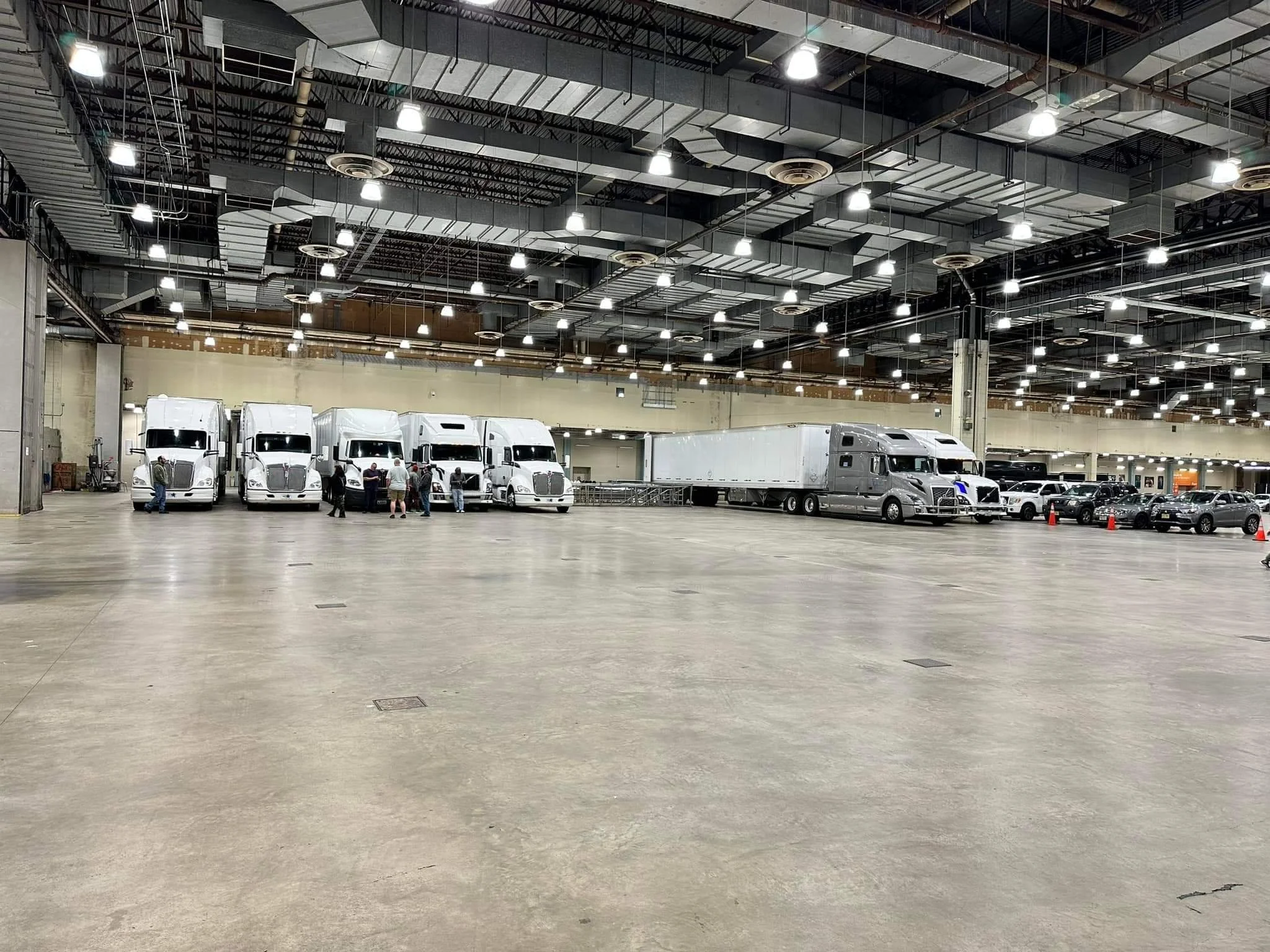 Interior of a large warehouse or logistics center with several white semi-trucks and trailers parked along the back wall. There are a few people near the trucks and several cars parked on the right side. The ceiling has many bright industrial lights.