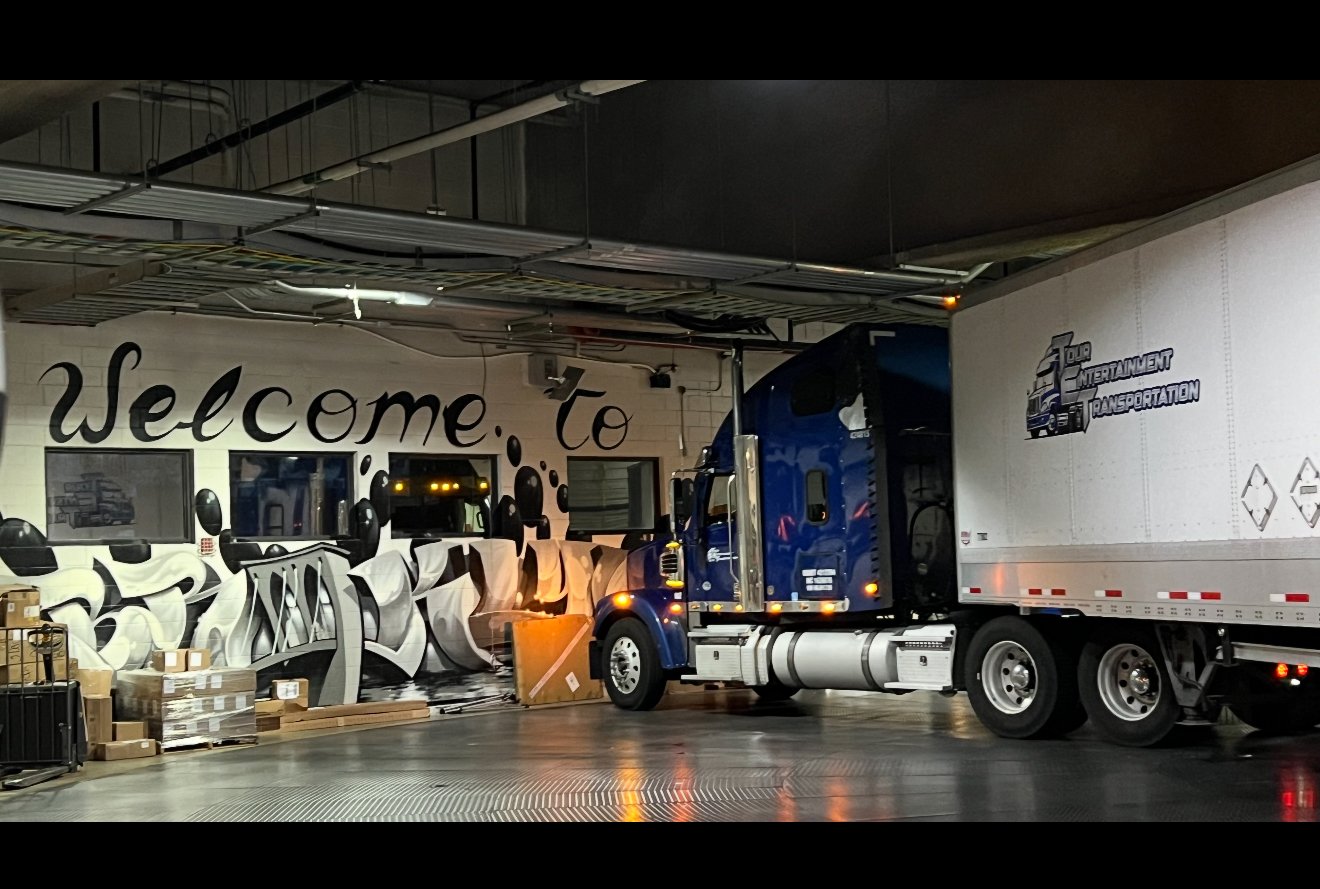 Inside a warehouse or loading dock area, with a large blue semi-truck parked near a wall with graffiti-style art that reads 'Welcome to'. A white container trailer is attached to the truck, and various boxes and packages are stacked nearby.