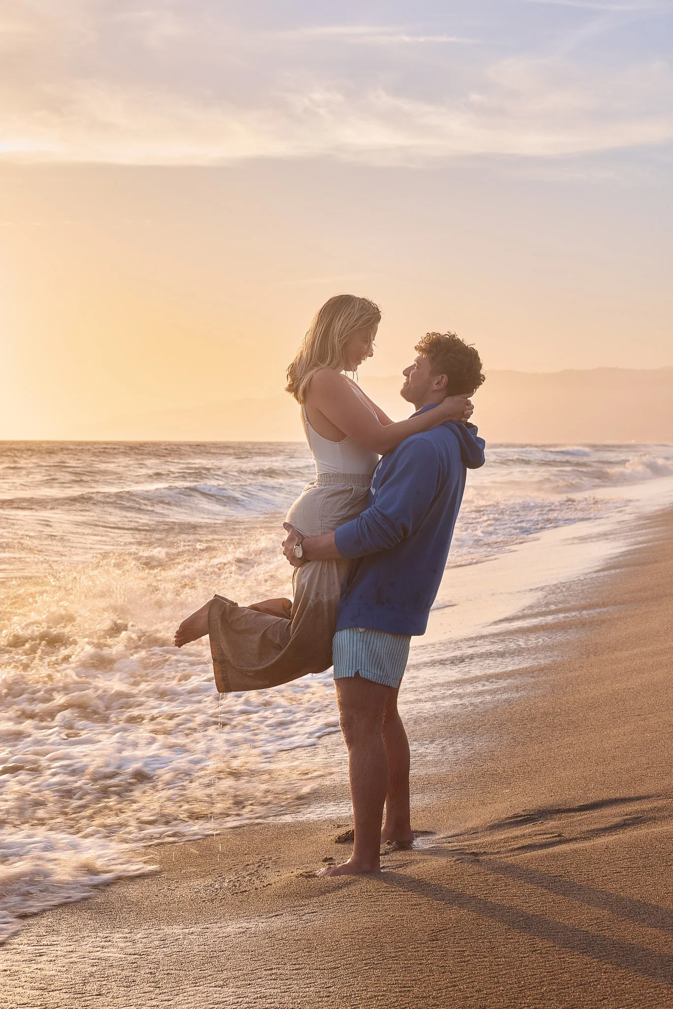 A couple on the beach during sunset, with the man lifting the woman in his arms as they look at each other, waves gently wash ashore in the background.