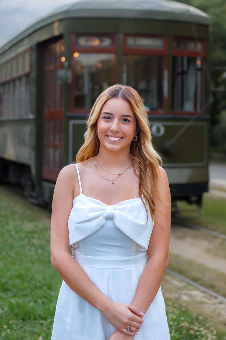 Woman in white dress smiling in front of vintage tram on grassy area.