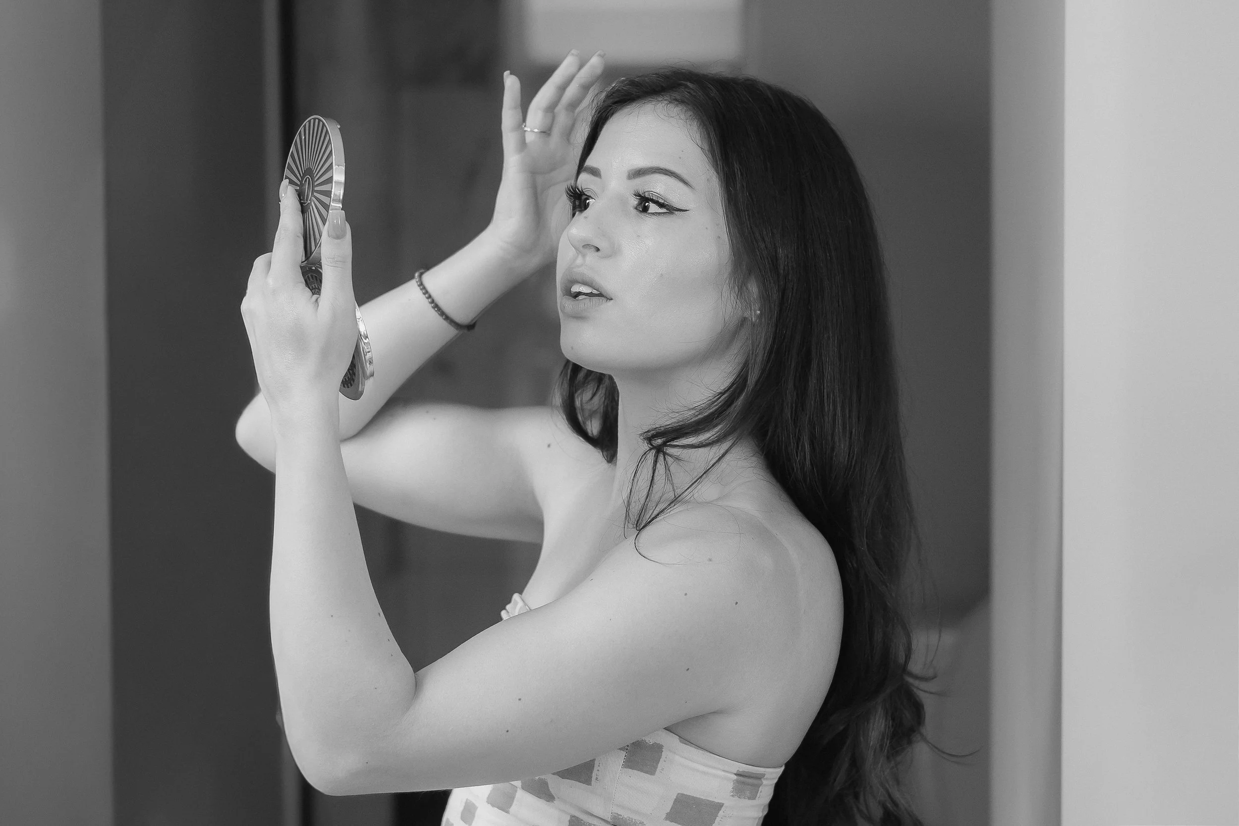 A woman with long dark hair is applying makeup to her face while looking into a handheld mirror.