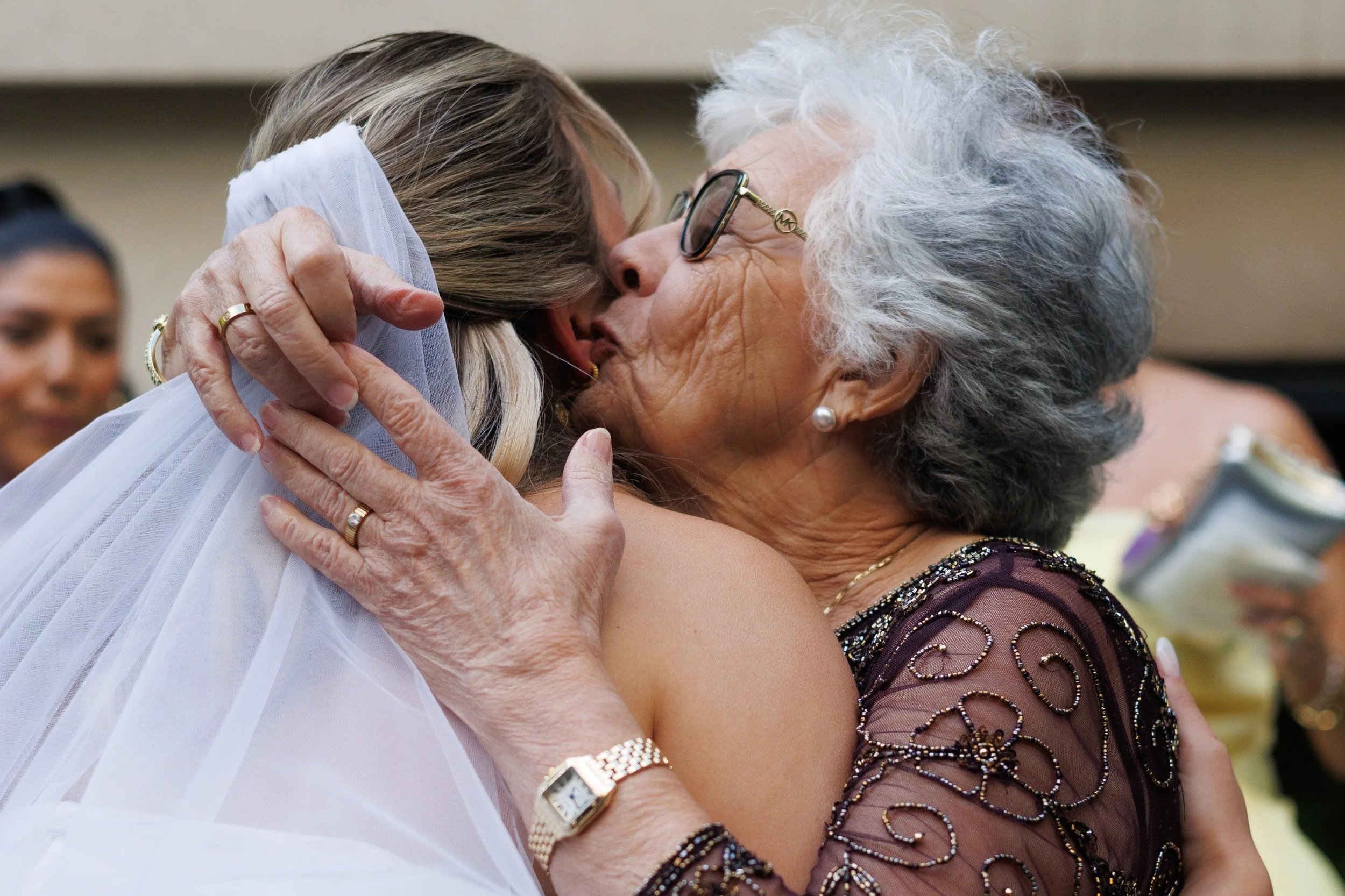 Chicago wedding photographer captures an elderly woman with gray hair and glasses hugging and kissing a bride wearing a wedding dress and veil, with a background of other guests.