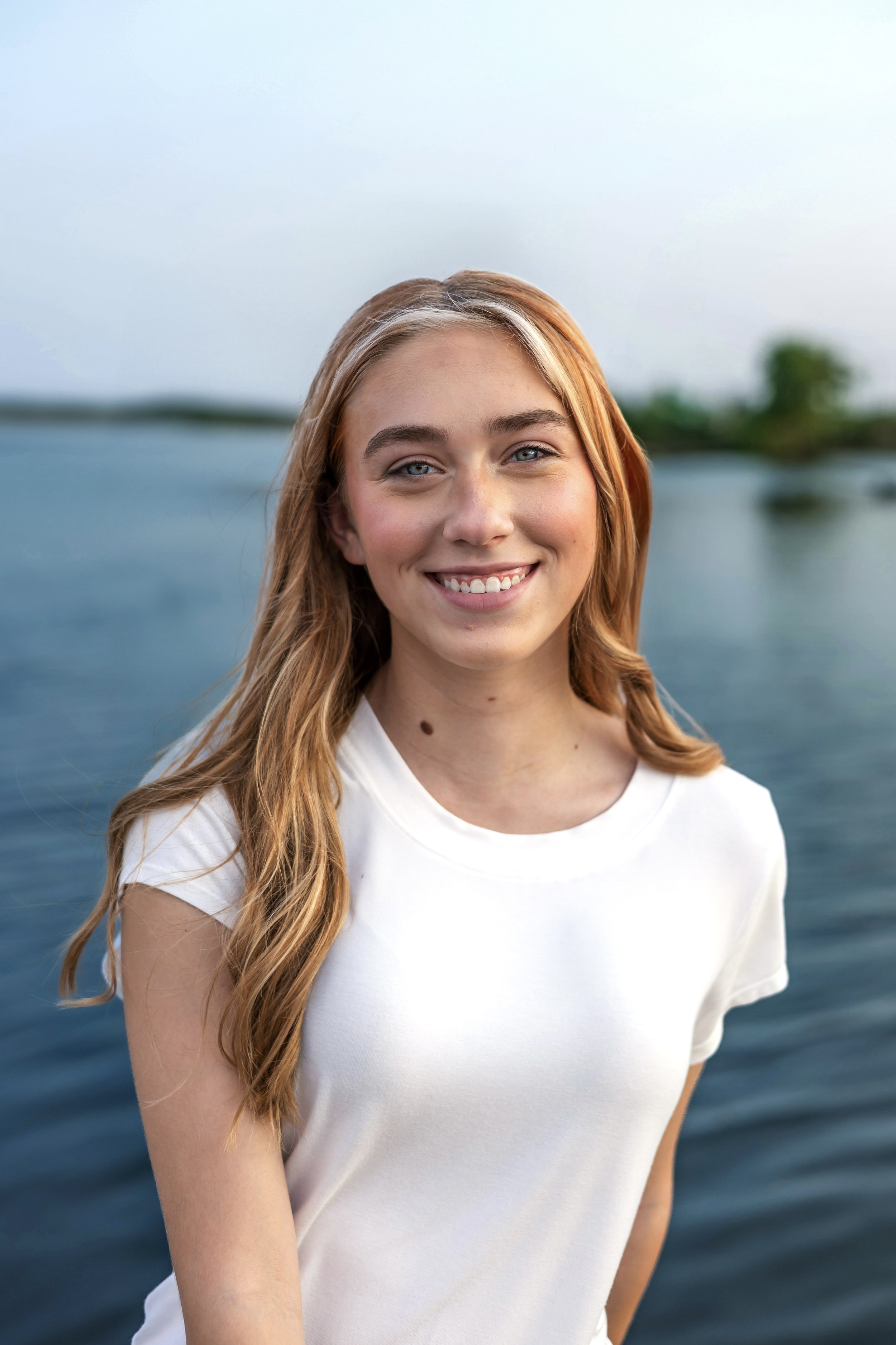 Smiling person with long hair by a lake, wearing a white T-shirt.
