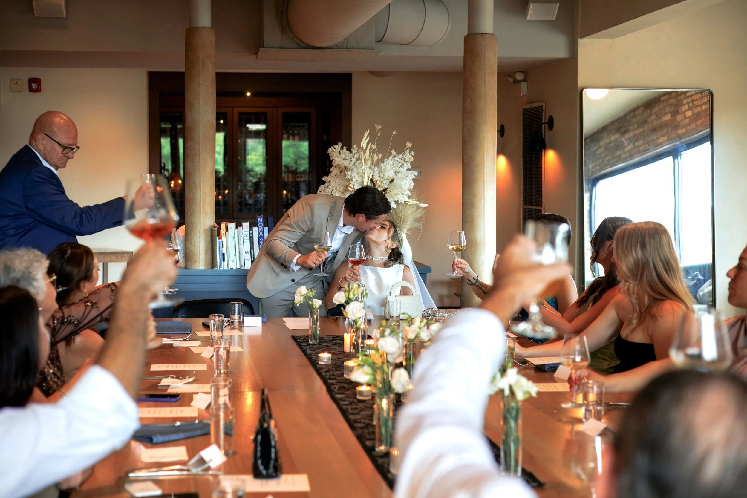 A wedding reception with a bride and groom sharing a kiss while raising glasses, surrounded by guests raising their glasses in a toast, in a decorated indoor setting with candles and flowers on the table, In the West Loop of Chicago