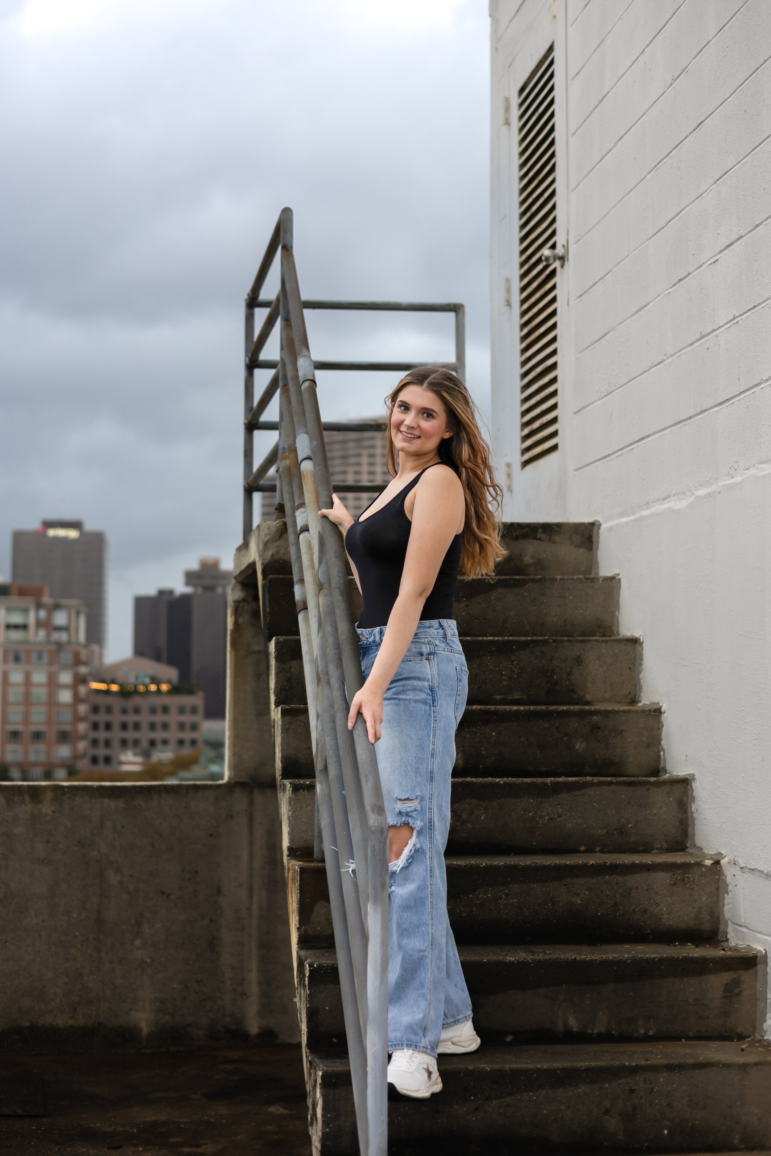 Woman in black tank top and ripped jeans standing on outdoor staircase with city skyline in background.