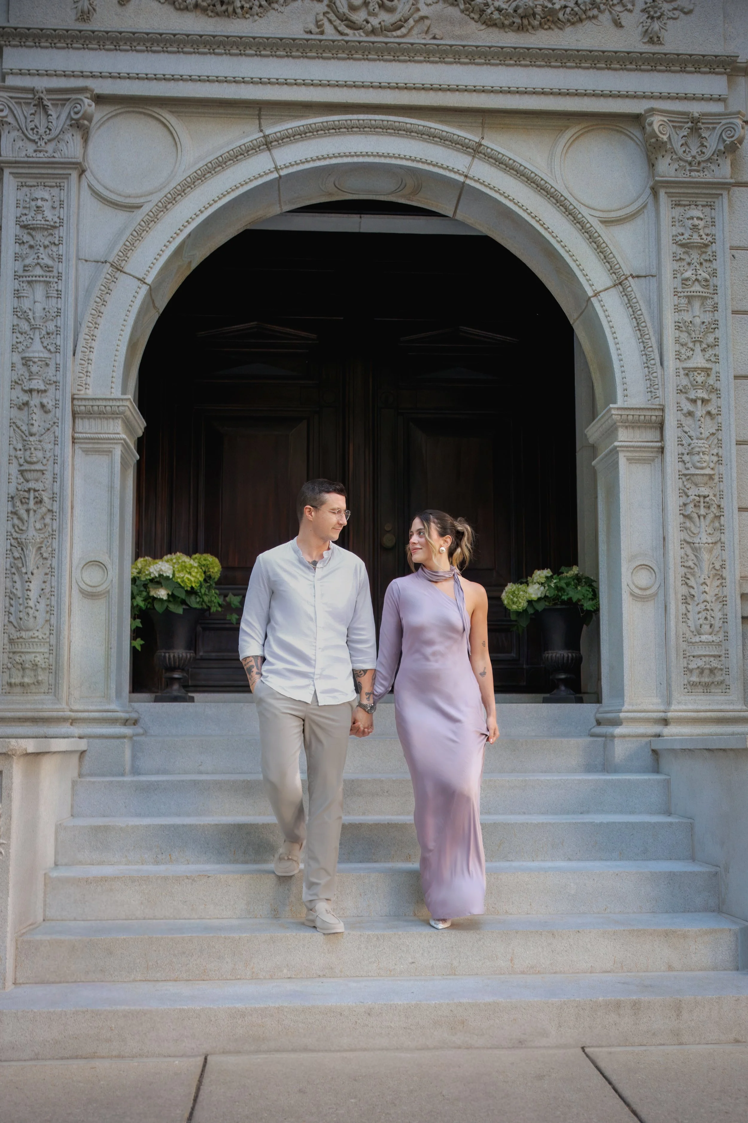 Engaged couple holding hands and walking down the steps of an ornate stone building while looking at each other, photographed during a romantic engagement session