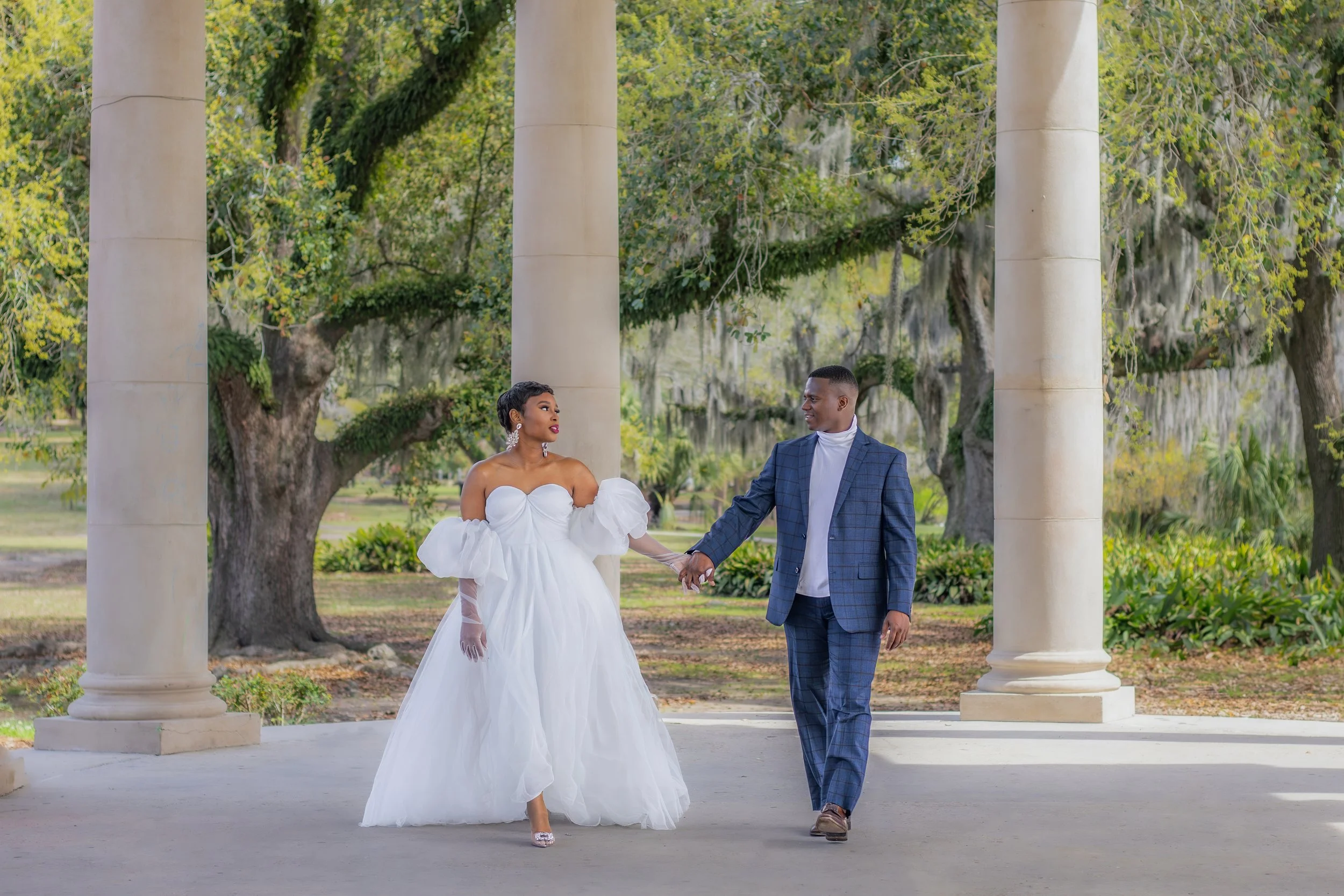 A couple holding hands, walking under stone columns in a park. The woman is wearing a white dress with ruffled sleeves, and the man is in a checkered suit with a white turtleneck. Surrounding them are large, mossy trees and greenery.
