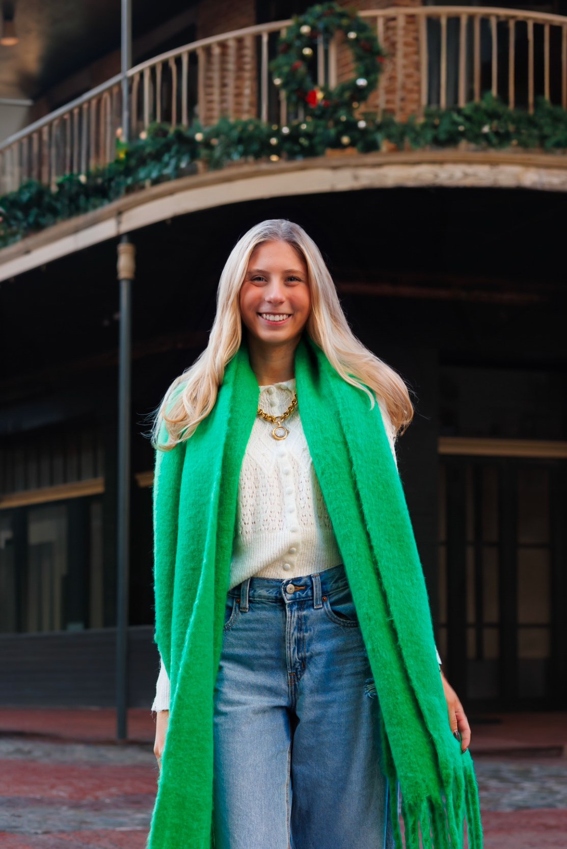 Woman in a white sweater and green scarf standing outdoors with festive garland and wreath in the background.