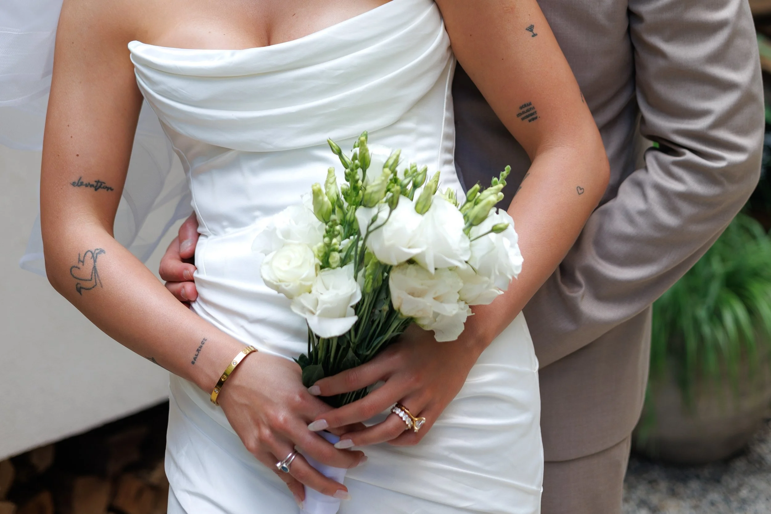 Close-up of a bride holding a bouquet of white flowers, with tattoos on her arms and rings on her fingers, coupled with a groom standing close behind her, partly visible.
Chicago wedding photographer