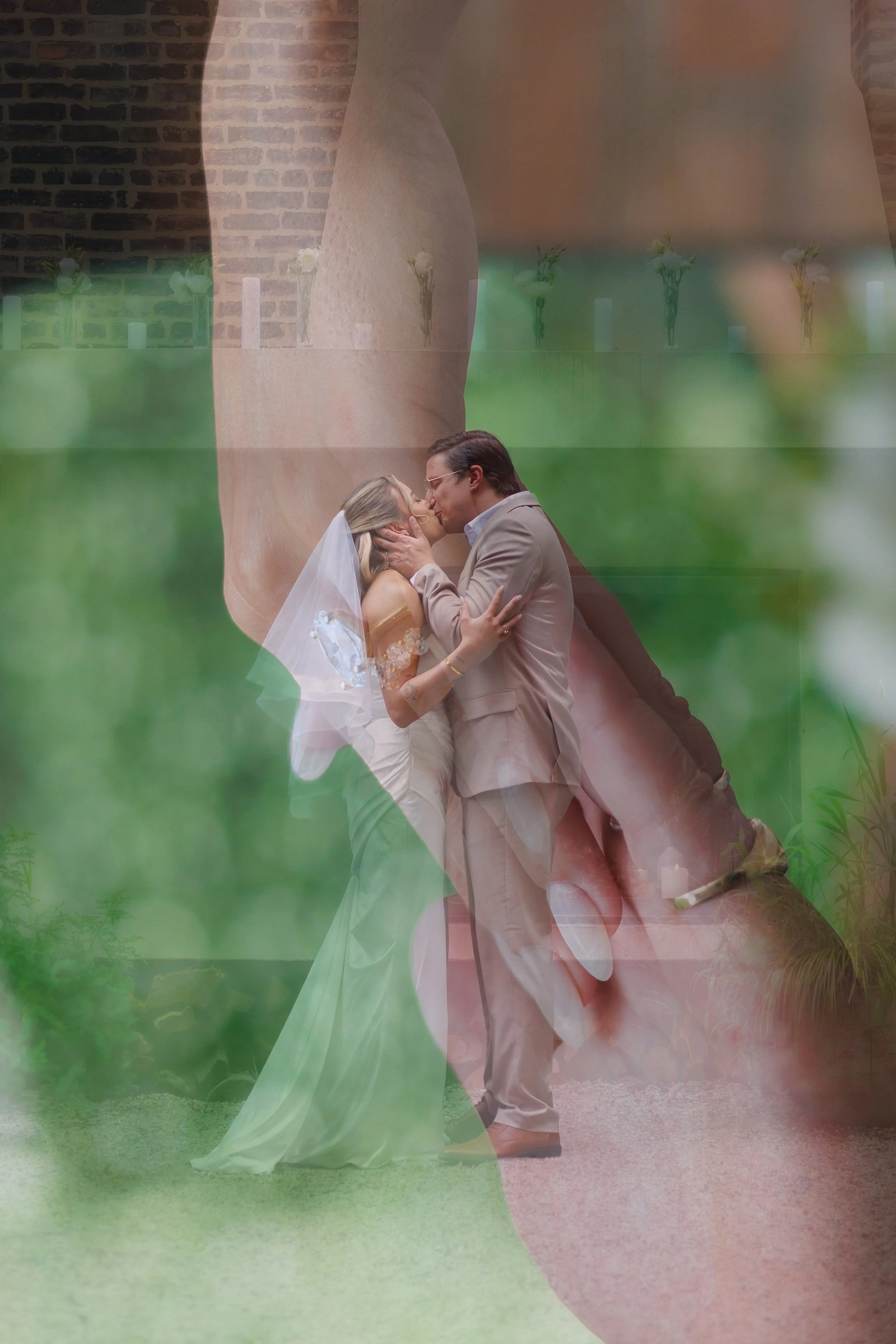 A couple in wedding attire sharing a kiss outdoors with a large, semi-transparent image of a woman in a bridal gown in the background.