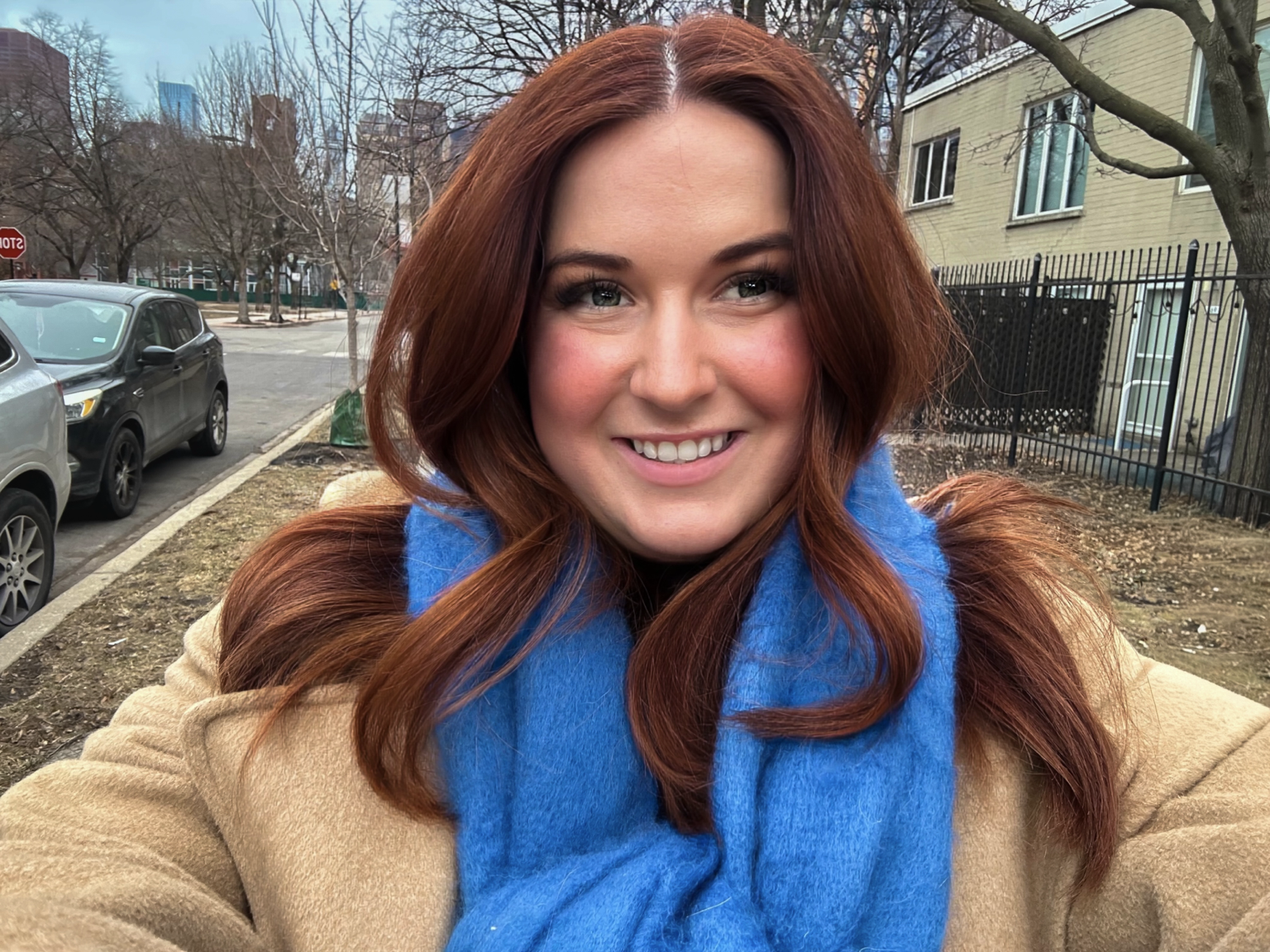 A woman with red hair smiling while taking a selfie outdoors on a sidewalk in a city.