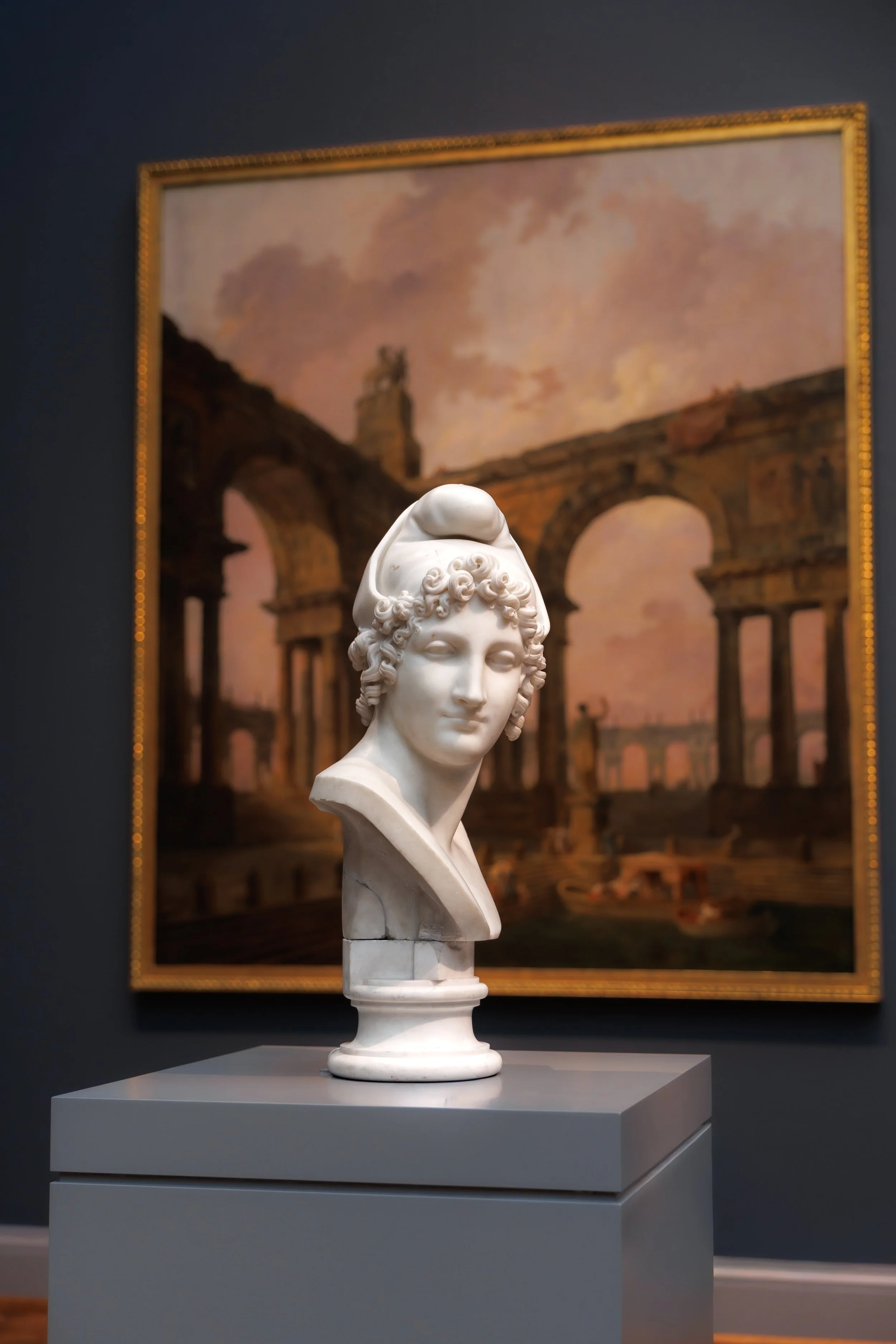 Marble bust of a woman with curly hair and head covering in an art gallery, with a landscape painting of ancient ruins in the background.