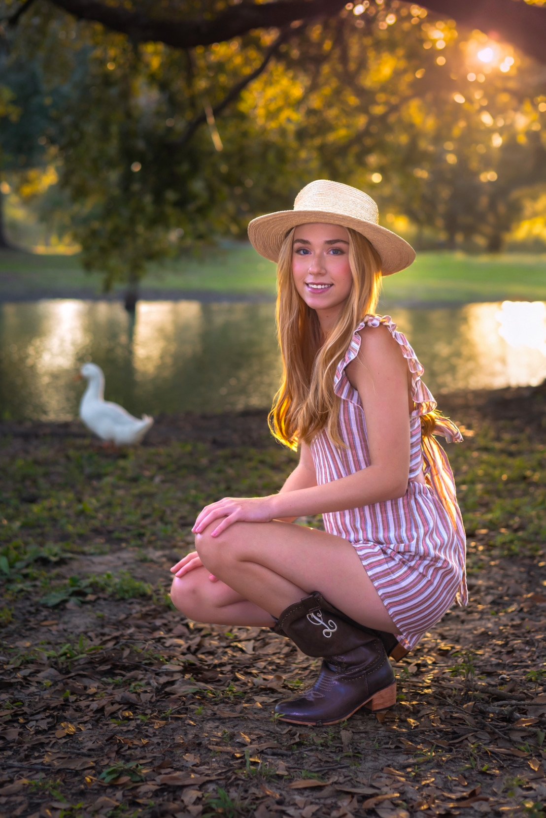 Young woman in a striped summer dress and straw hat crouching near a pond with a duck, surrounded by lush greenery and dappled sunlight.