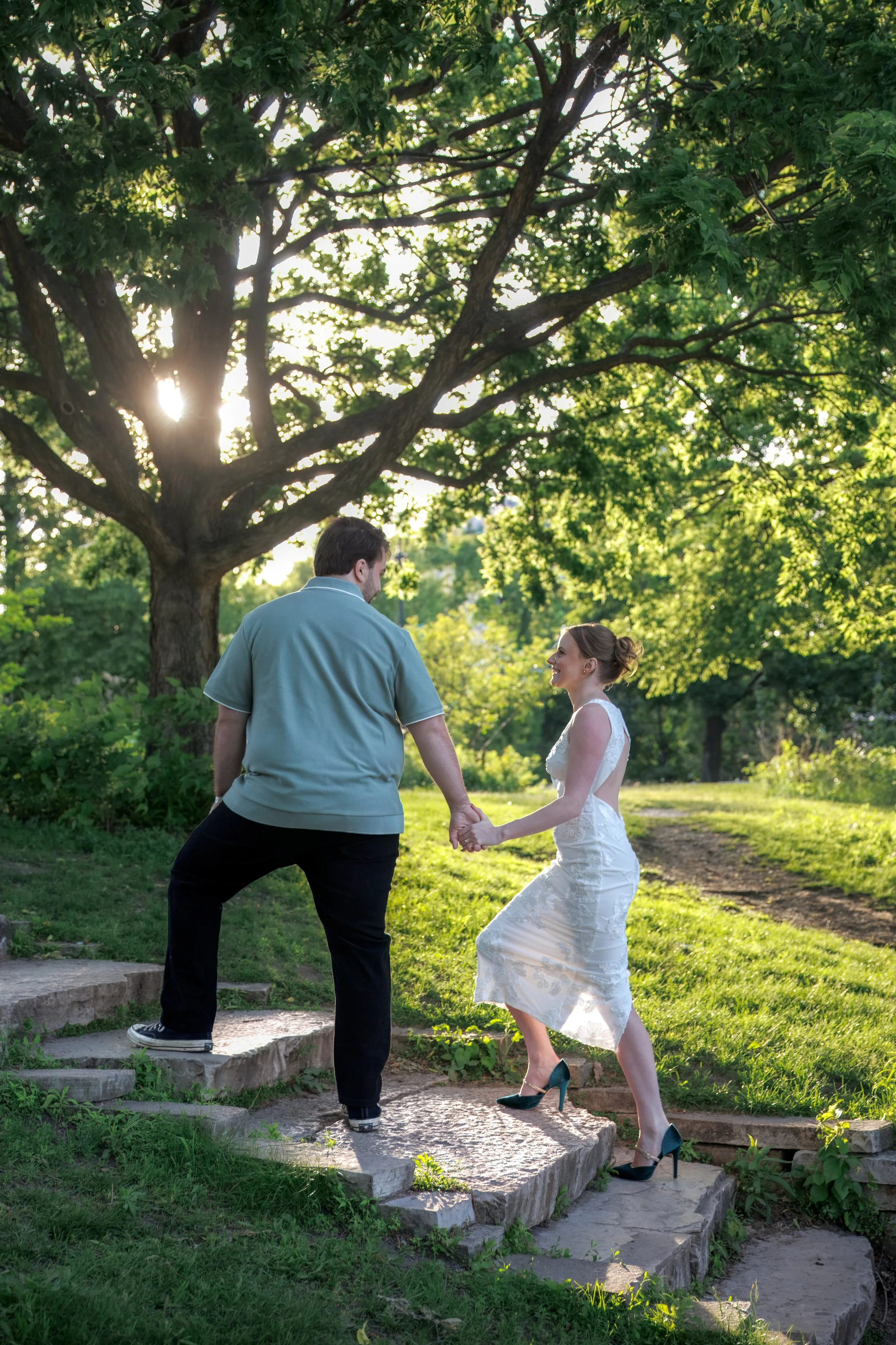 a couple walking up the stairs with lush trees behind them and soft light peeking out.