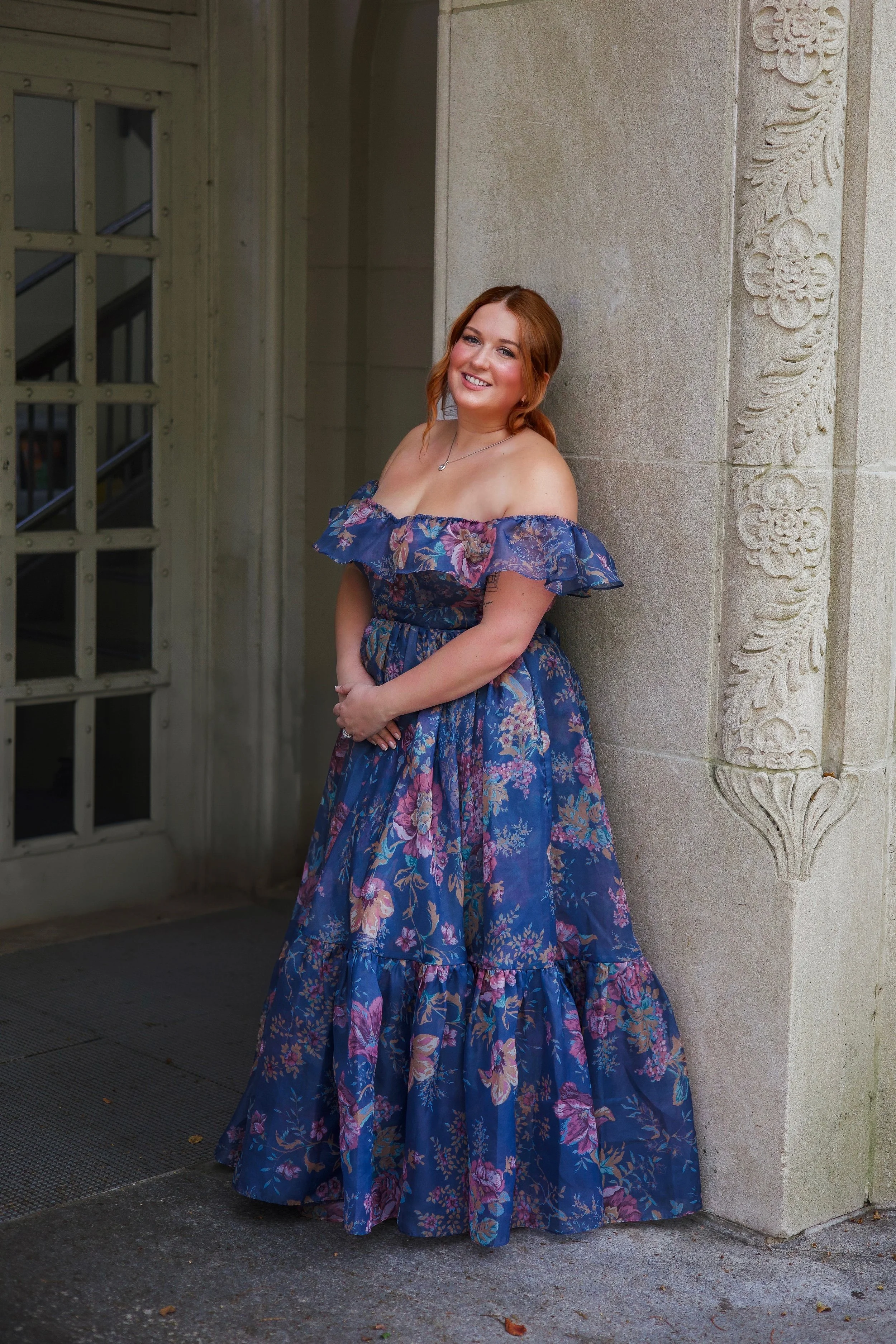 A woman with red hair in a floral blue off-shoulder dress standing next to a decorative stone wall with carved floral patterns, smiling at the camera at the University of Chicago