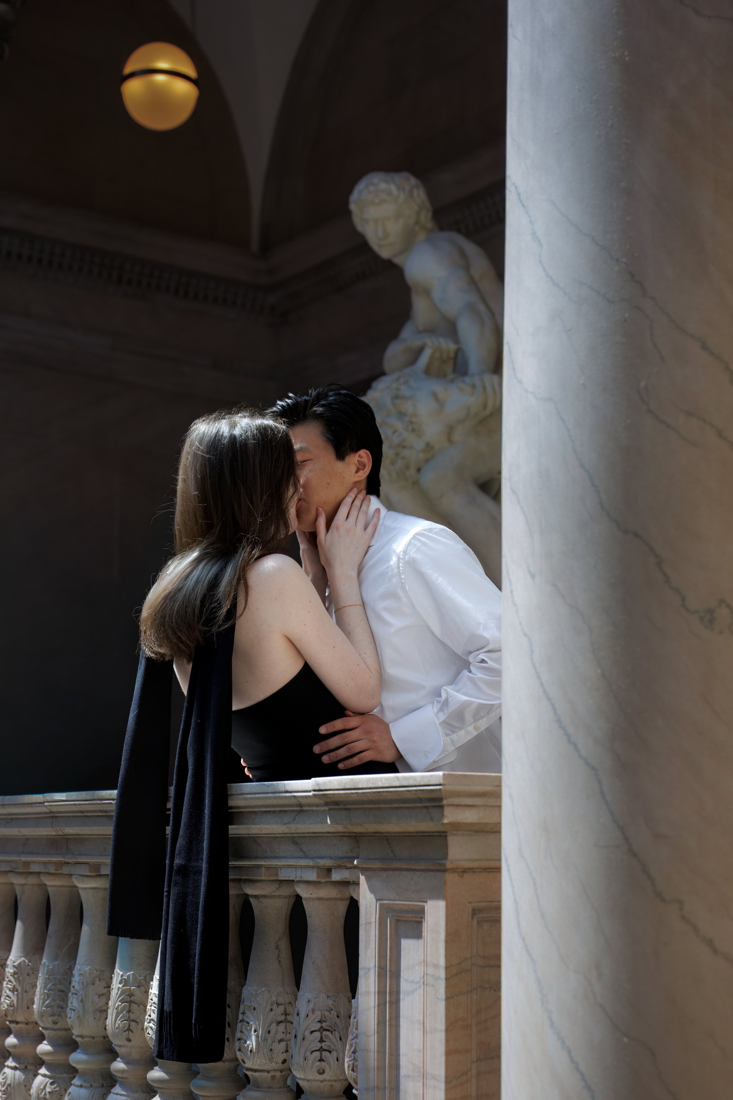 Couple kissing on a marble balcony inside the Art Institute of Chicago, framed by stone columns and classical architecture, with a sculpture watching in the background.